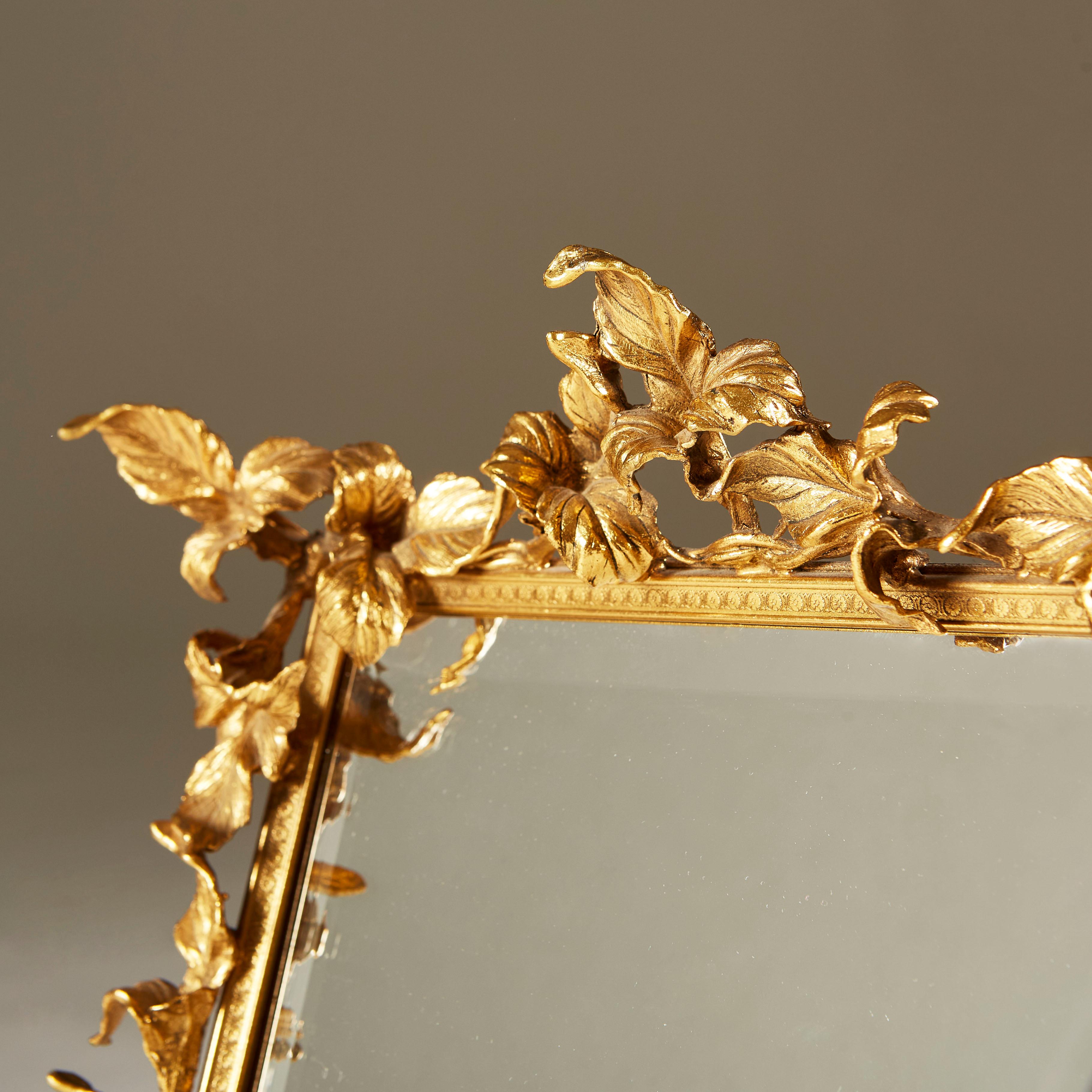 Dekorativer goldfarbener Schminktischspiegel mit kreuzförmig gemustertem Rahmen und aufwändiger Blattverzierung. Sitzt auf Kugelfüßen. Samtrücken und Rückenstrebe. Spiegel mit abgeschrägter Kante.


