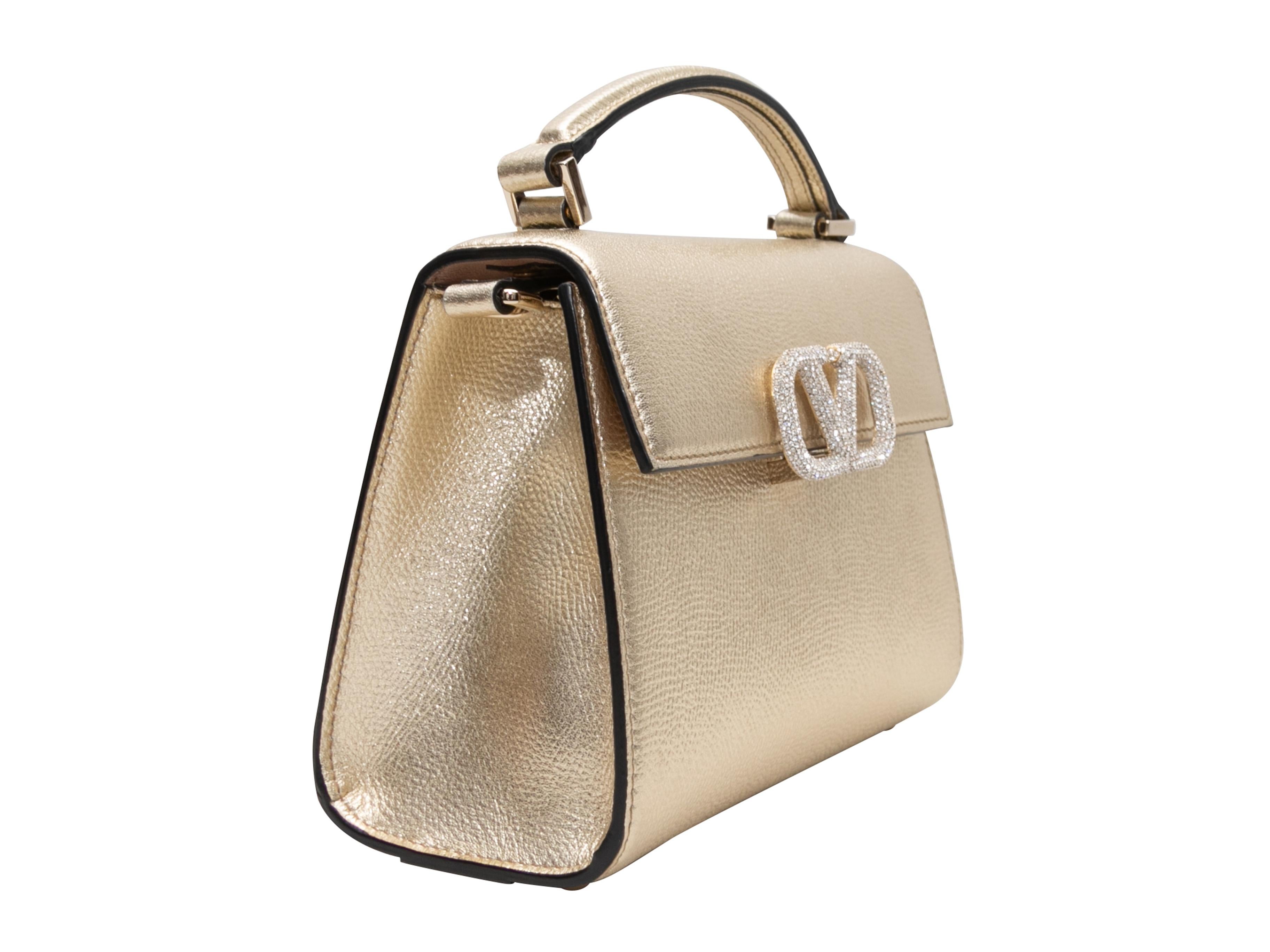 Product Details: Gold Valentino Mini Vsling Crystal Logo Bag. The Mini Vsling bag features a metallic leather body, single top handle, optional crossbody strap, and front crystal-embellished logo flap closure. 7.5