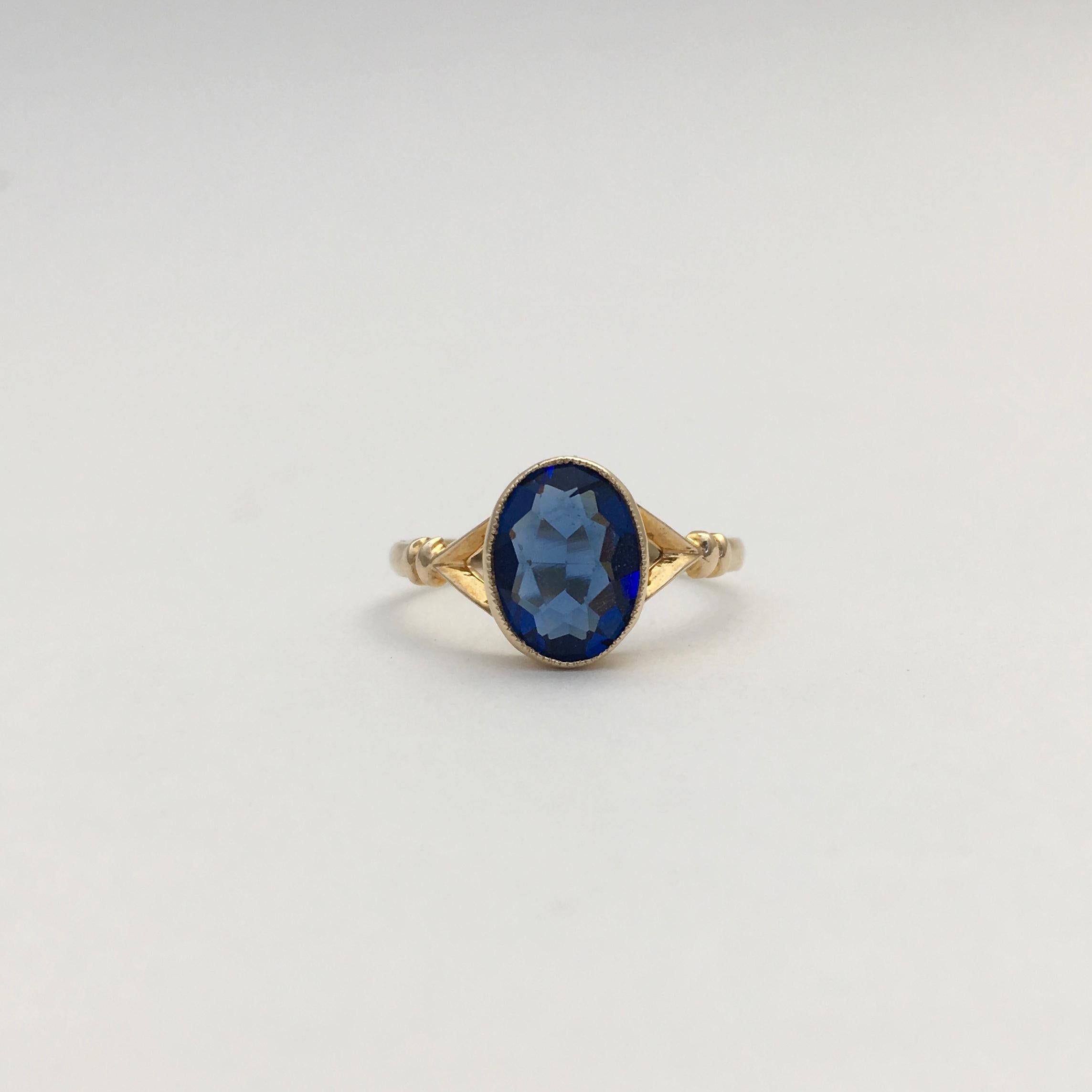 This pretty little 9ct gold ring is a striking cobalt blue colour. The dainty style dates it to the early to mid 20th century. A delicate millegrain setting surrounds the synthetic oval stone, which is made to resemble a sapphire. The stone measures