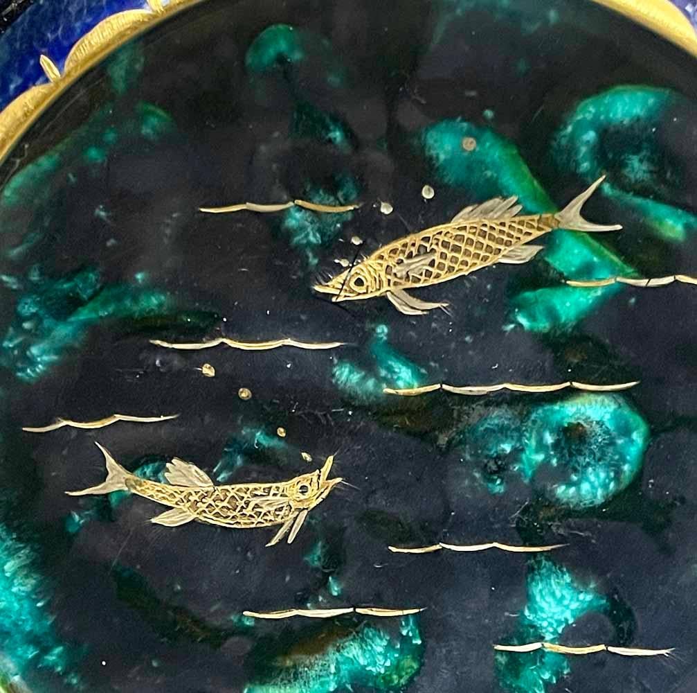 Lovely and unique, this footed bowl or dish depicts two golden fish, incised into the clay and then glazed in gold, swimming in a dark blue sea flecked with deep green.  The artist was Auguste Heiligenstein, known for creating pieces with exotic