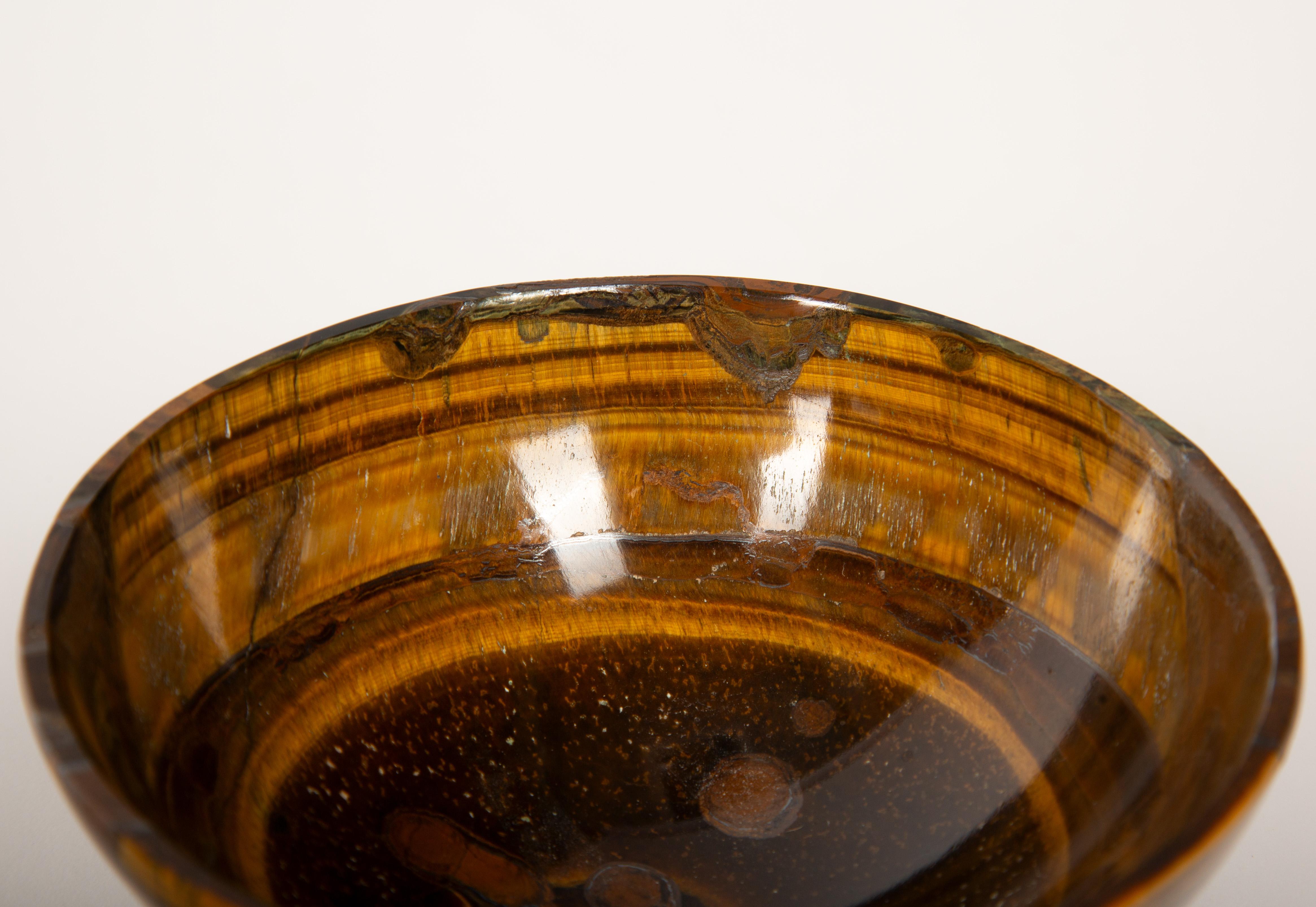 Questa piccola ciotola di grande effetto è scolpita nell'occhio di tigre lucidato, una gemma semipreziosa apprezzata per la sua lucentezza setosa e per le sue bande color oro-marrone che cambiano alla luce. Con un diametro di circa 4,25 pollici, è