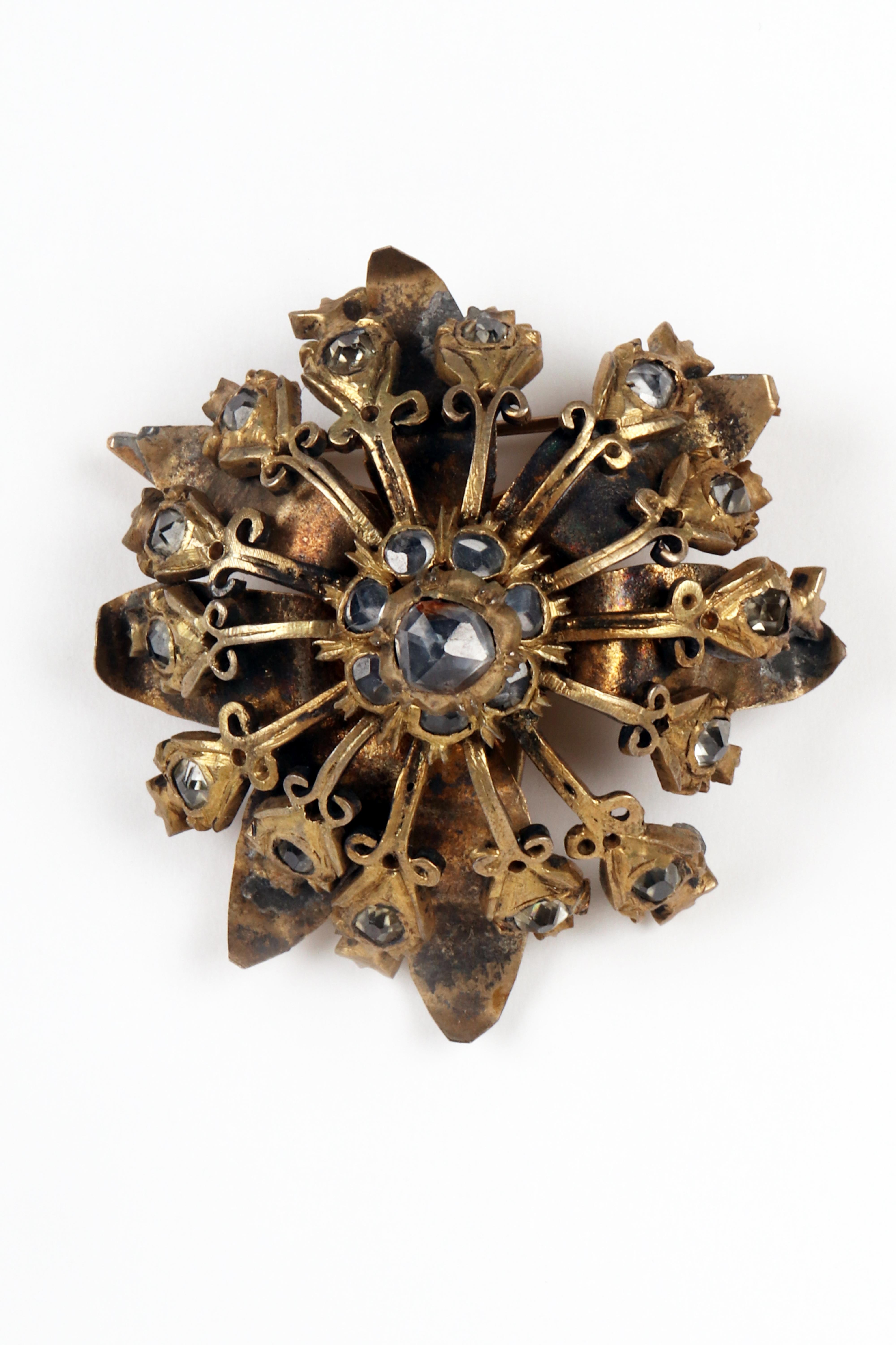 Button-parade brooch in golden metal and diamonds from India for the English market. The base is made up of a continuous laminar body element that opens into petals with twisted tips at the center of which is hinged a first radial pattern of