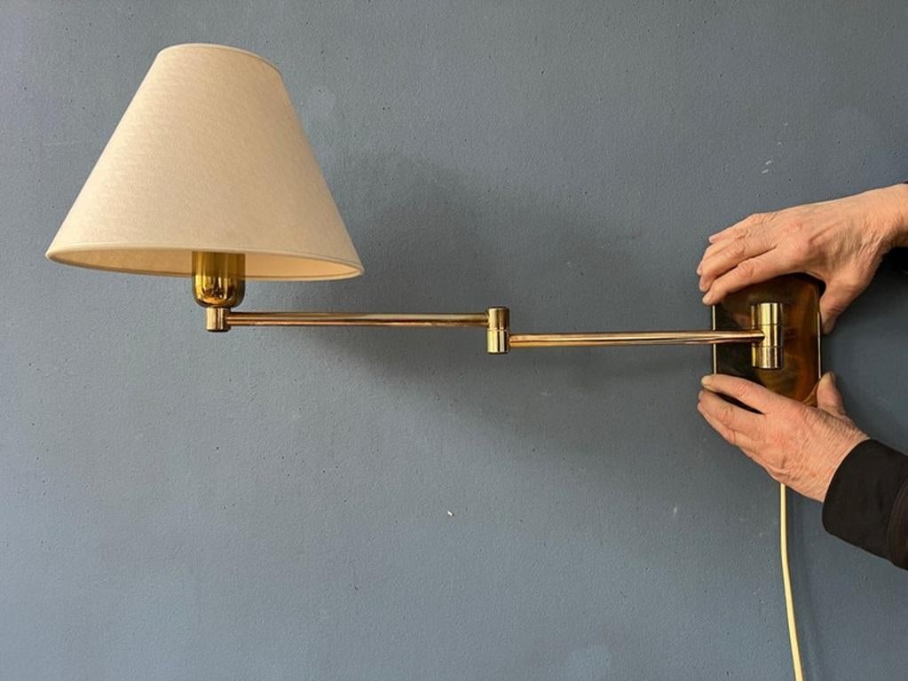 Lámpara de pared de brazo oscilante Hollywood Regency con estructura dorada y pantalla beige. La longitud y la posición de la lámpara pueden ajustarse fácilmente con el mecanismo basculante. La lámpara requiere una bombilla E27/26 (estándar) y
