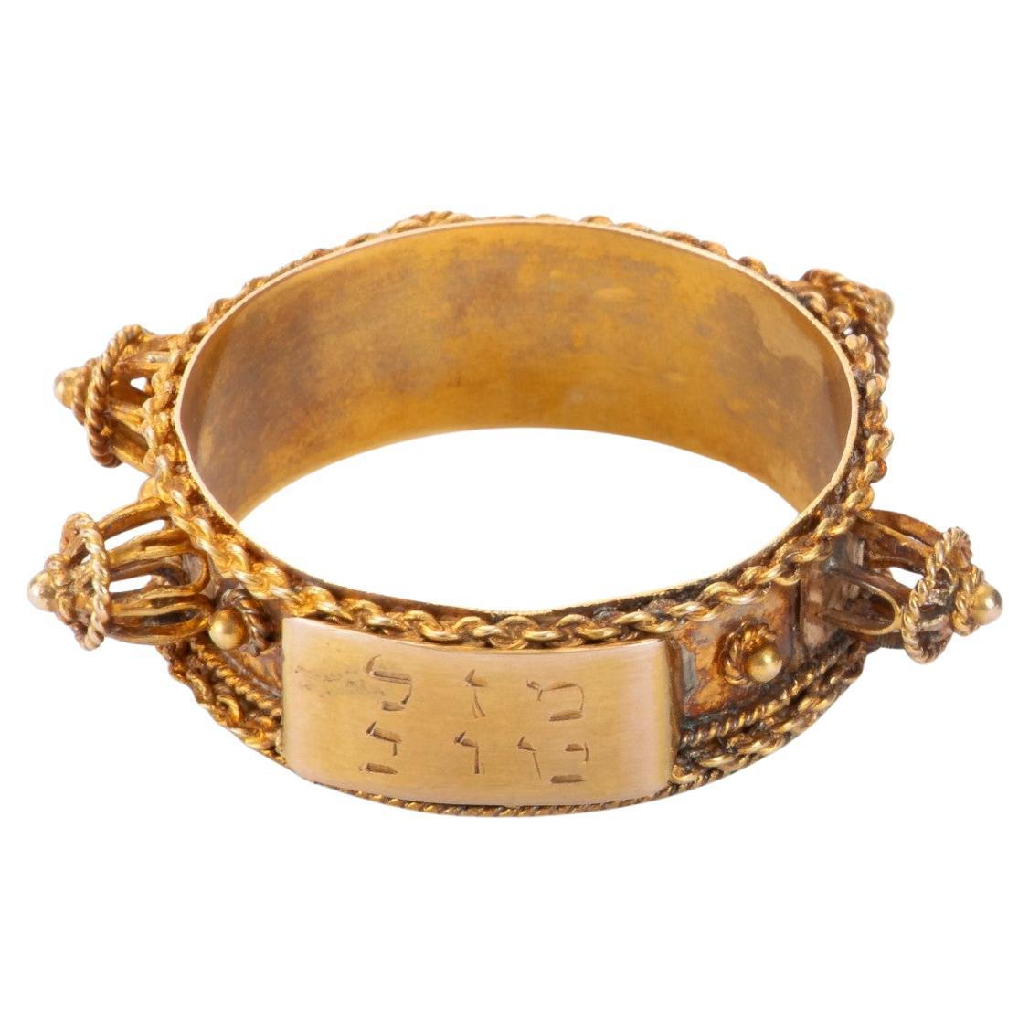 Un anello nuziale ebraico elegante e storicamente evocativo, realizzato interamente in oro e delicatamente ornato da una minuziosa lavorazione in filigrana. Catene d'oro sottili e intrecciate a mano circondano i bordi esterni dell'anello,