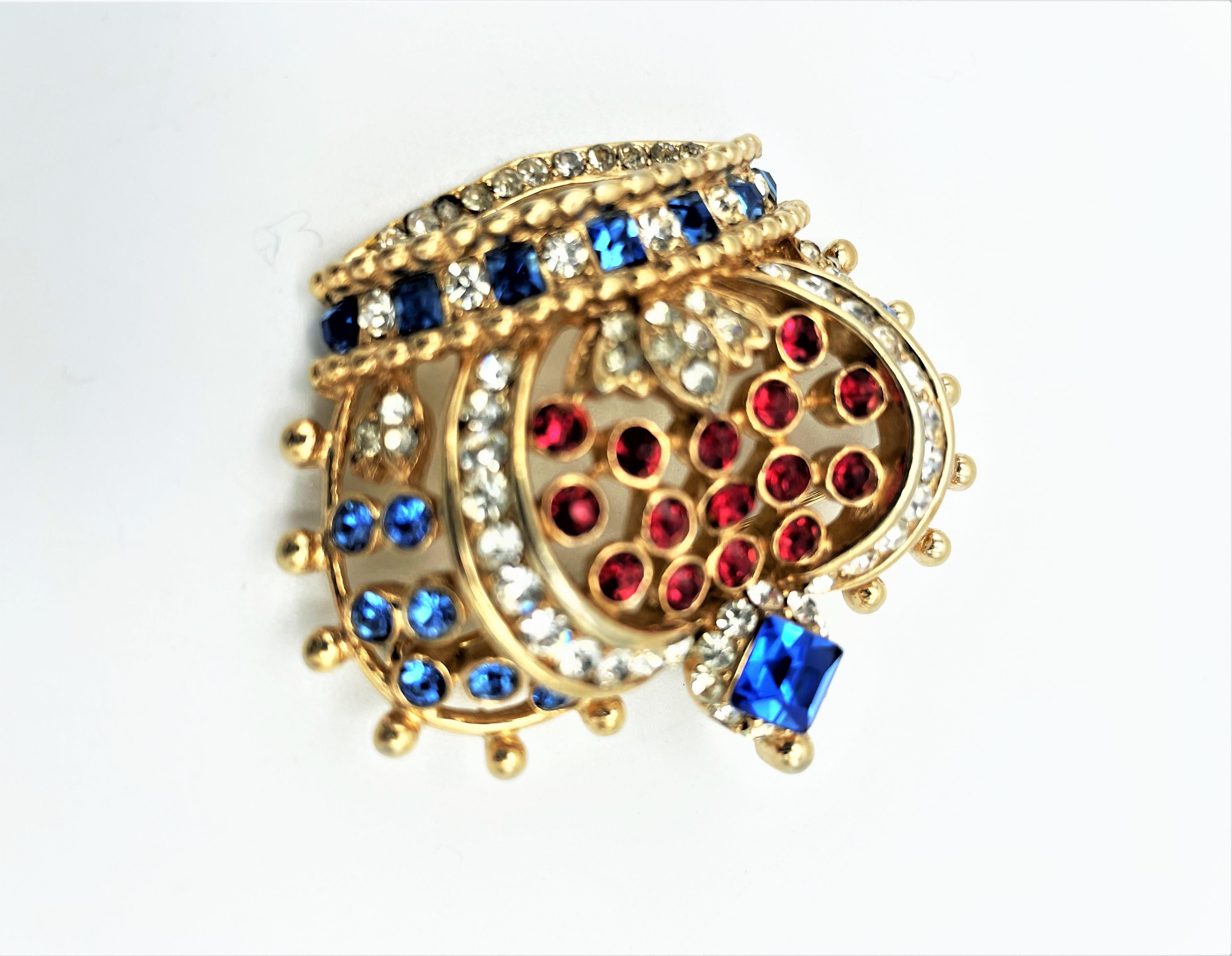 A royal crown set with red, blue and clear rhinestones. The brooch is crowned with a sapphire colored rectangular polished rhinestone. Gold plated bass metal USA around 1950s.

Measurement: Width 5 cm, Height 4,5 cm, Deep 1,5 cm, the sapphire stone