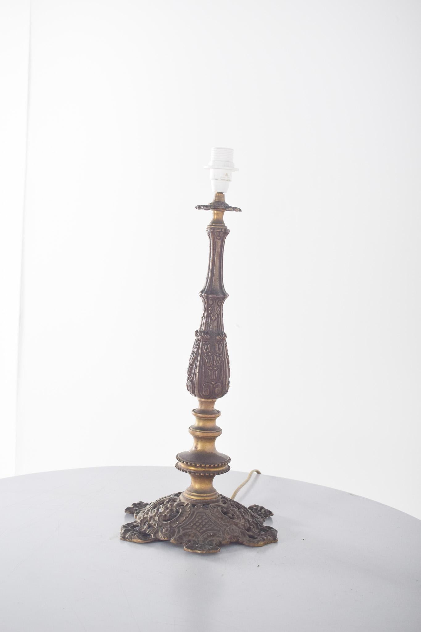 Table lamp in gilded metal.  Finished in aged bronze, it has a base decorated with floral details and arabesques in relief, harking back to the Baroque style.
The body of the lamp is elongated and carved with decorative patterns in high relief,