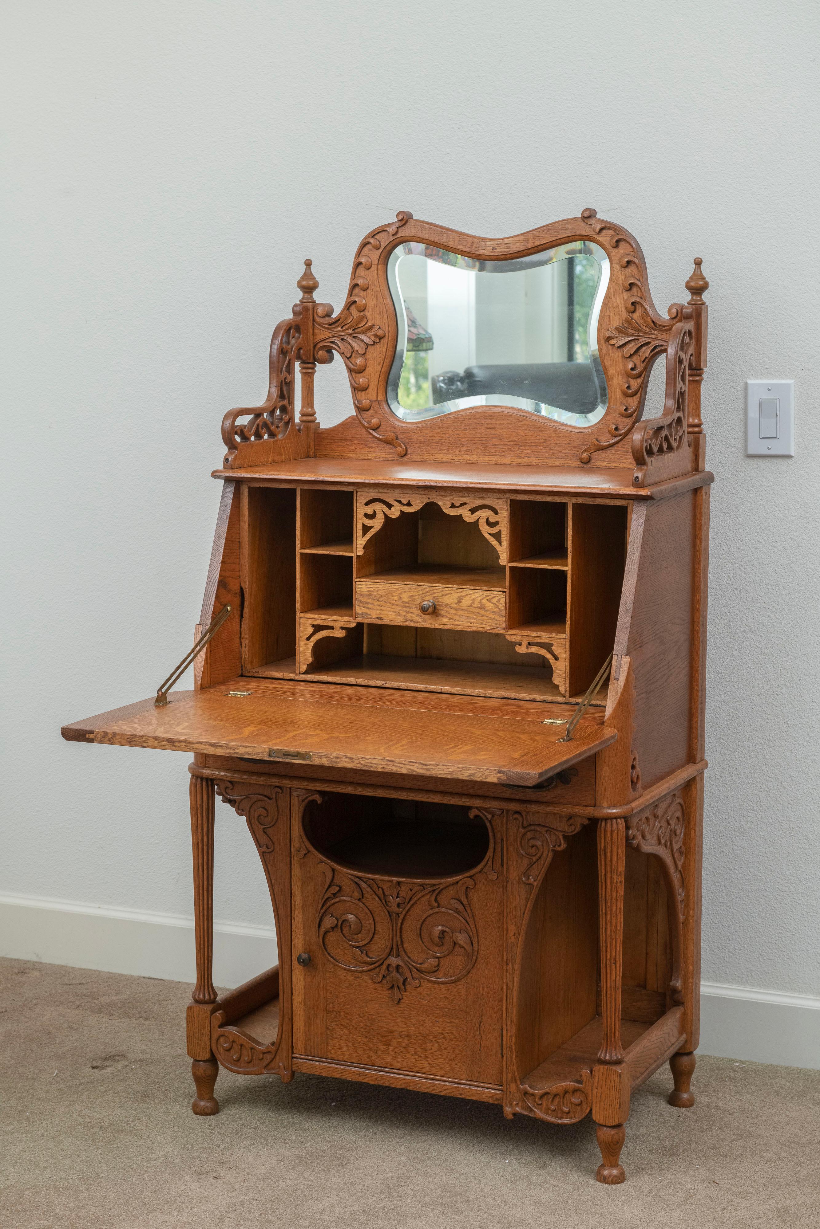 This dazzling golden oak secretary/desk is feast for the eyes. The leaf drops down for use, and opens up displaying a little drawer and a bunch of cubby holes. Above the desk area is a shelf w/carved sides and, a curvy beveled mirror, w/carving as