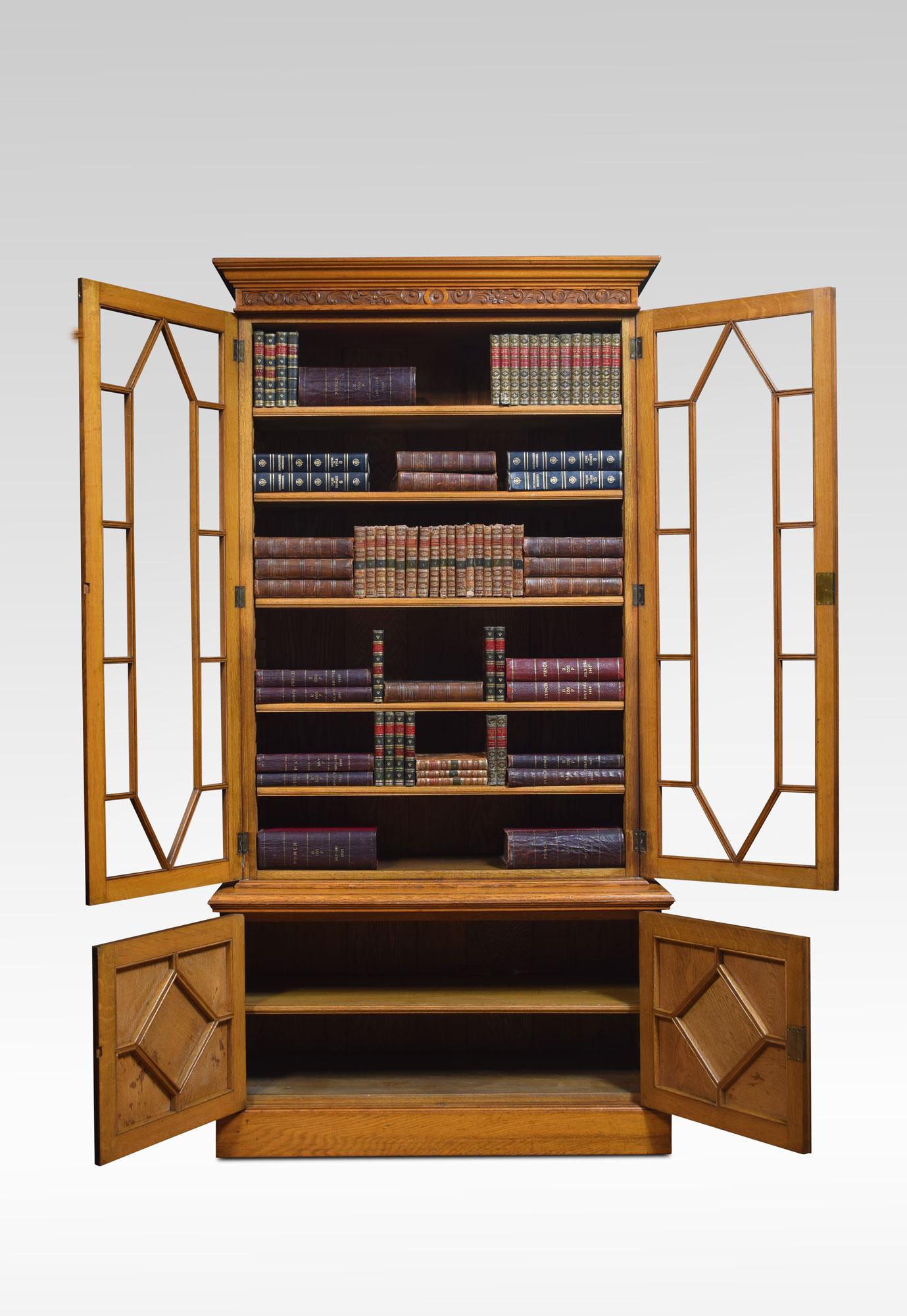 Golden oak bookcase, the moulded cornice and carved frieze above two large astragal bevelled glazed doors opening to reveal an adjustable shelved interior. To the base fitted with a pair of geometric moulded doors. All raised up on plinth
