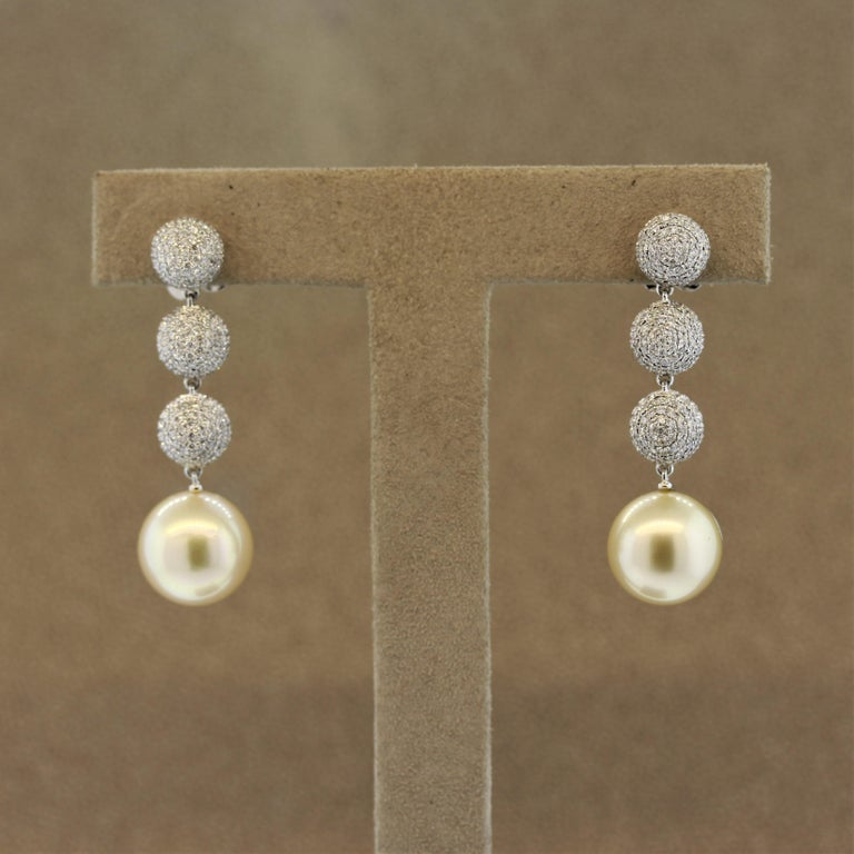 A luxurious pair of diamond drop earrings featuring 2 golden south sea pearls measuring 12.5 millimeters. They are suspended below 3 diamond set gold spheres adding length and movement to the piece. Made in 18k white gold, feel like a star in these