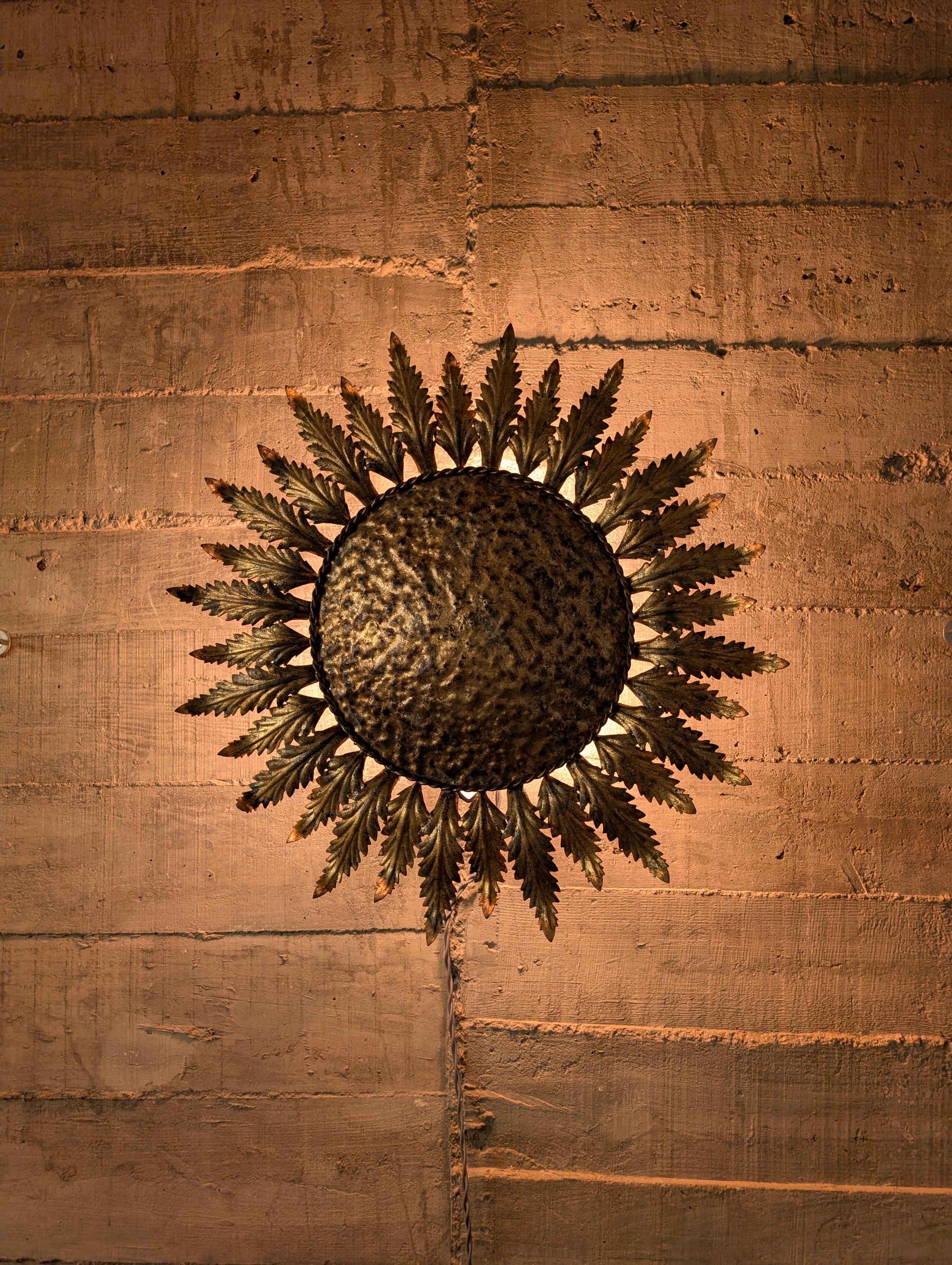 Preciosa lámpara de mediados de siglo en forma de sol con rayos de hoja en metal dorado, que puede utilizarse como lámpara de techo o como aplique de pared. Presenta tres puntos de luz que resaltan su encantadora silueta y su impresionante pátina