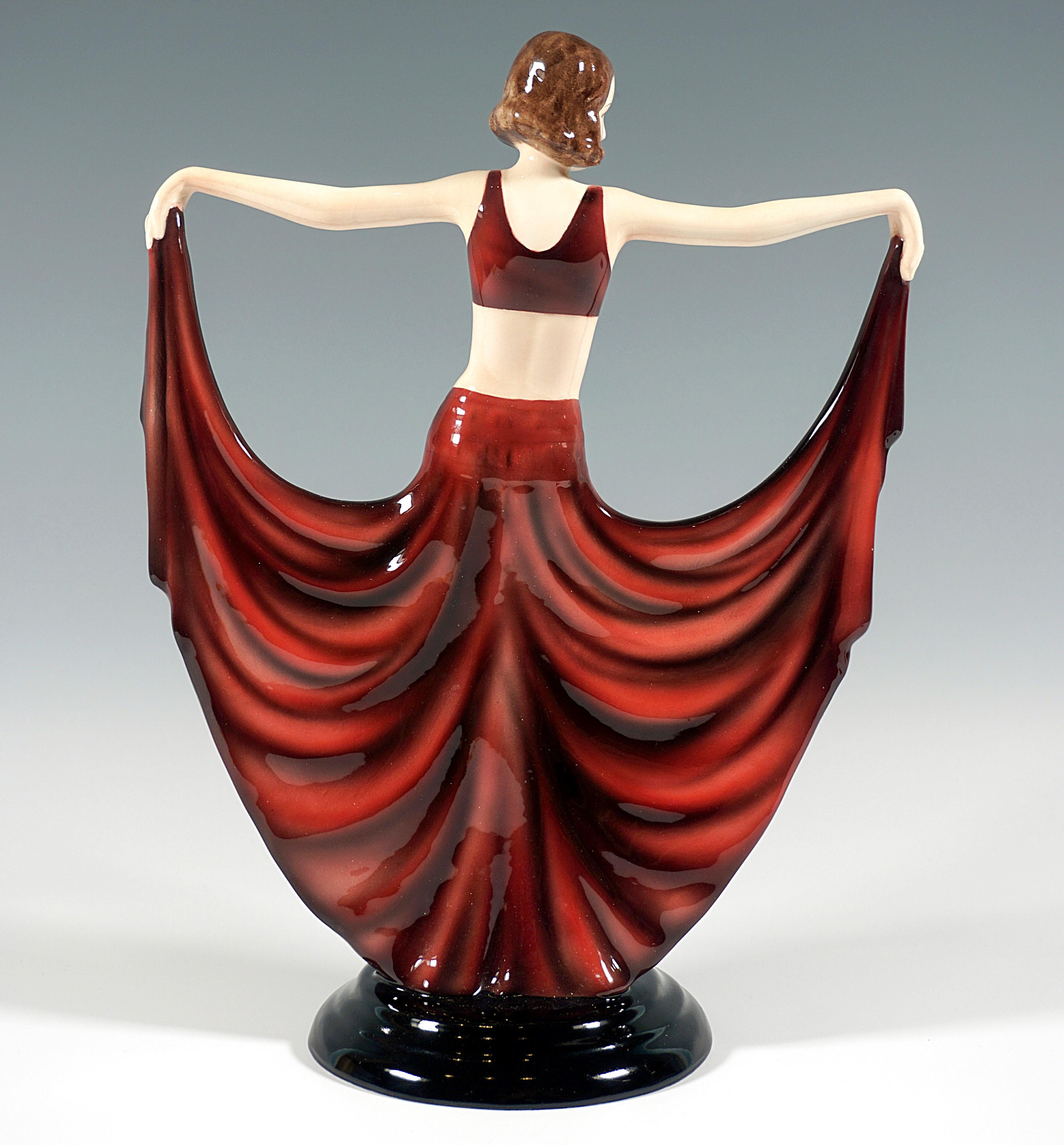 Danseuse à la pose gracieuse, vêtue d'un costume à dominante rouge : bustier et jupe longue, large et très fendue, avec des décorations bleues et jaunes en forme d'ailes de papillon. Elle tient son costume avec les bras tendus de chaque côté, la