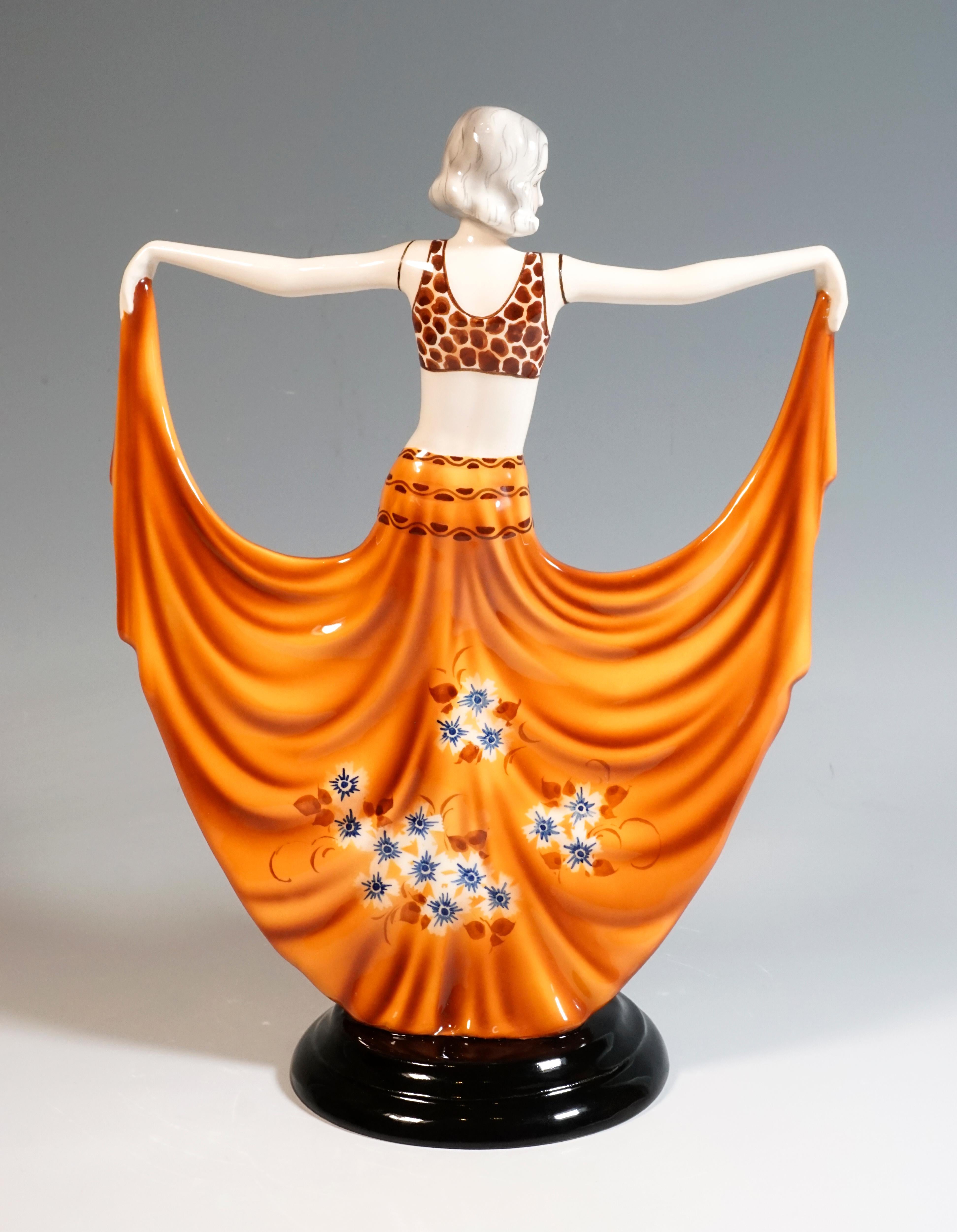 Gracefully posing dancer in orange costume: bustier and floor-length, wide, front slit skirt with blue floral decoration, holding it up like a butterfly on both sides with outstretched arms.
On a black, stepped, fluted round base.

Signed STEPHAN