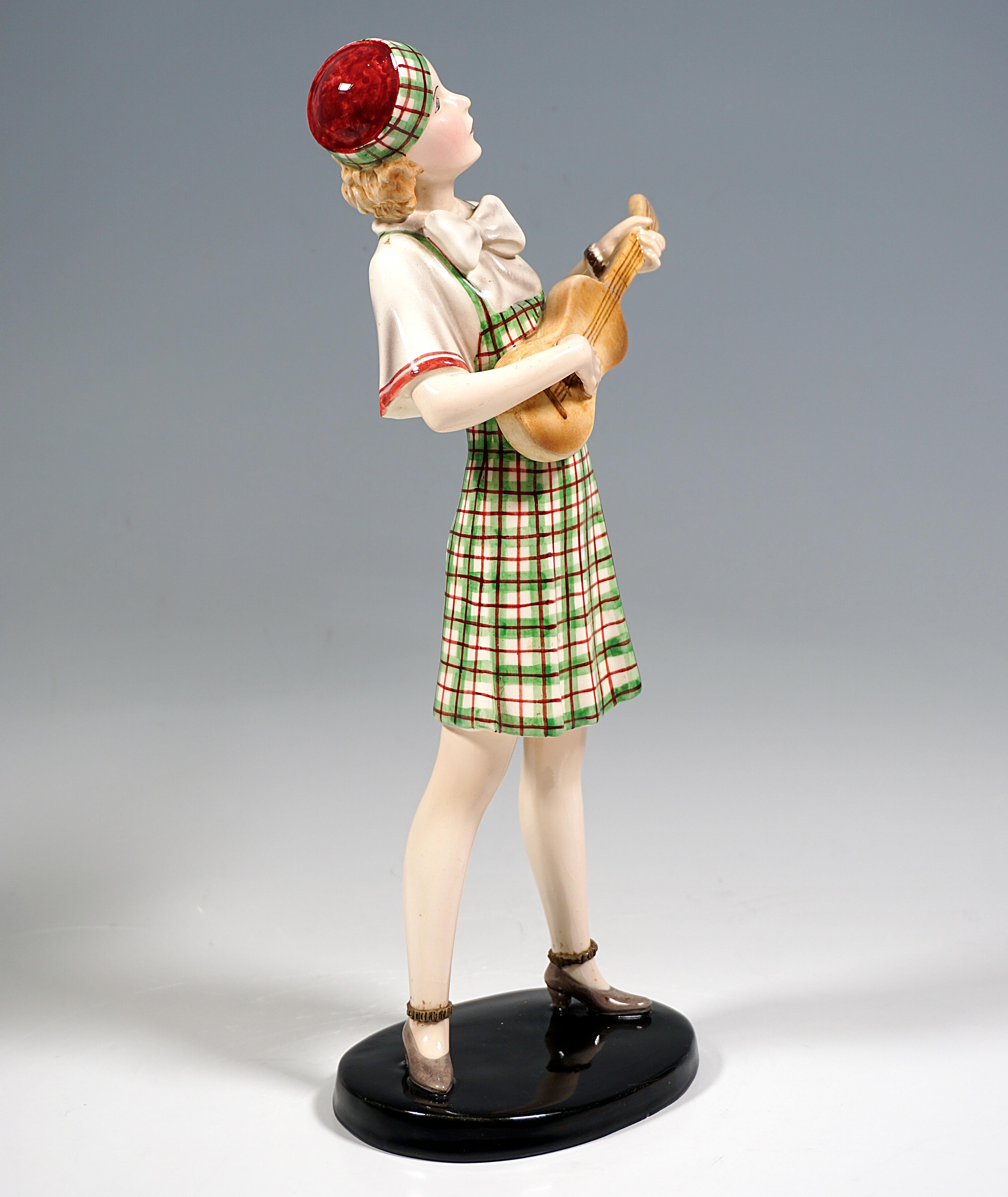 Very rare Goldscheider Vienna Ceramic Figurine of the 1930s:
Standing young lady with blond hair in beige-green-red checkered dress over blouse with large mesh and wide red hemmed short sleeves, on hair a cap matching the dress, playing ukulele and