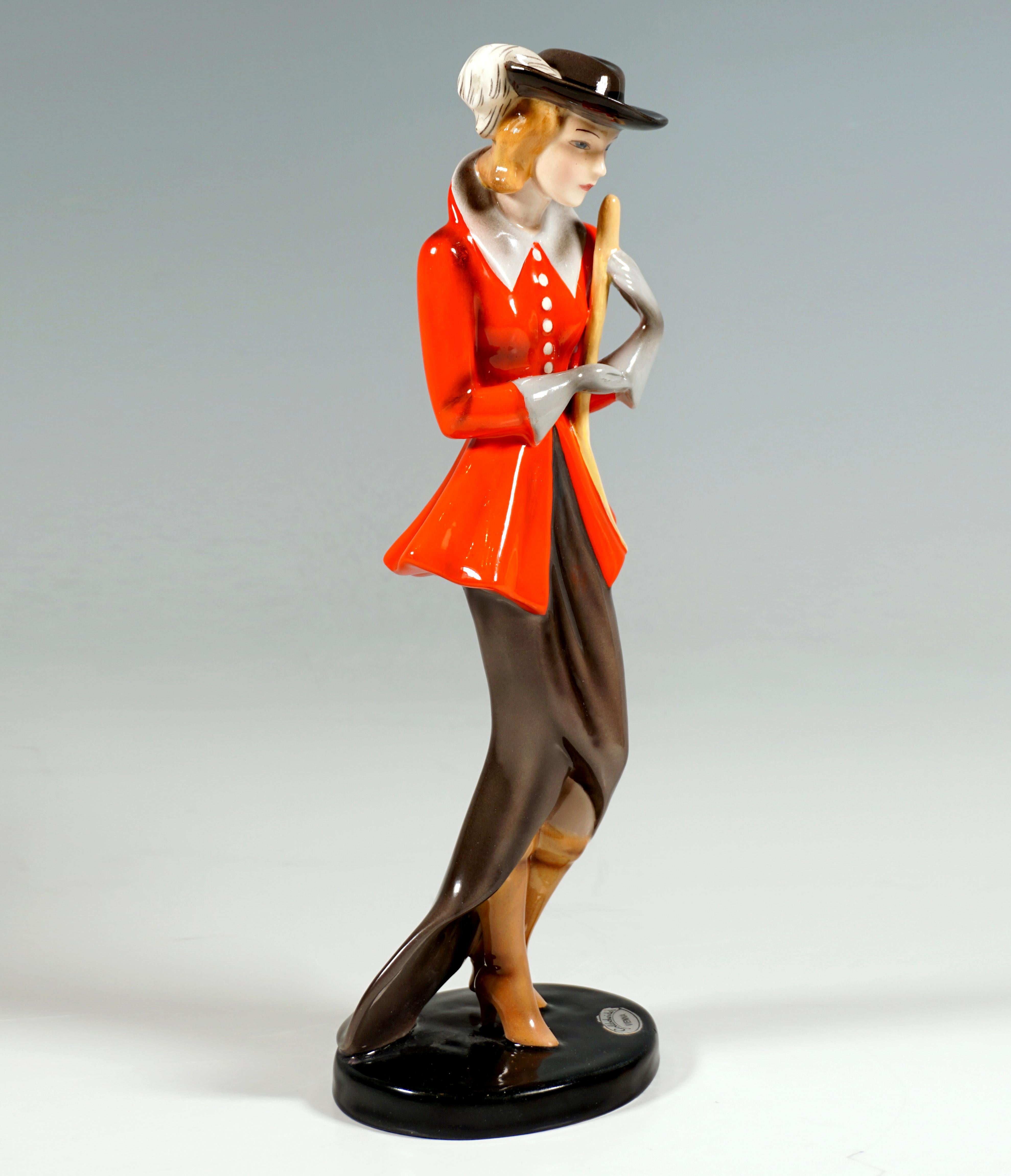 Very Rare Goldscheider Vienna ceramic figure of the 1930s:
Standing, elegant lady with a dark feather hat, in a long, dark skirt and a red, tailored jacket with a high, light-grey collar and matching gloves, holding a riding crop in both hands.
On