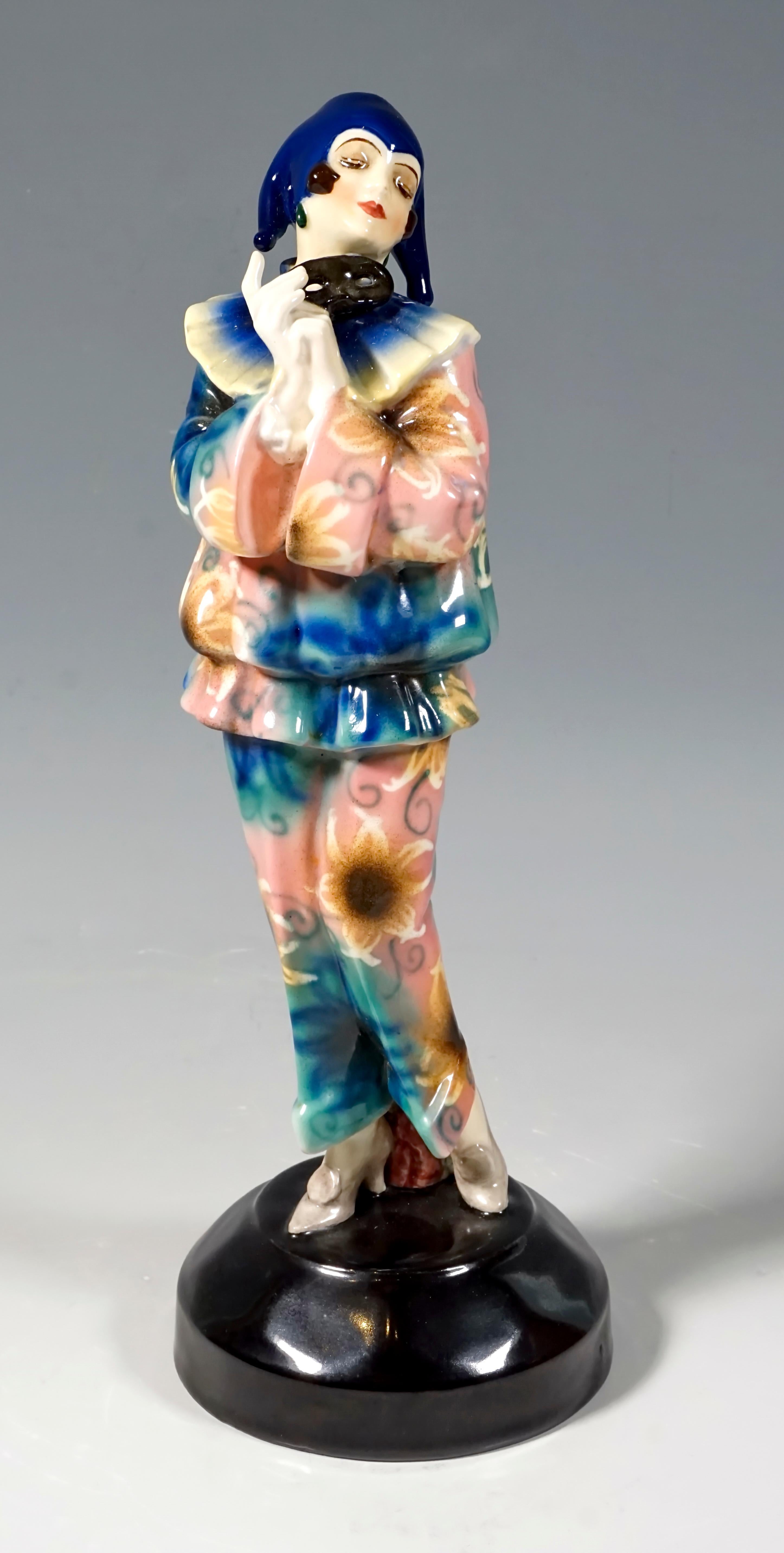 Finest Viennese ceramic art from the 1920s:
The standing young lady holds a black mask in front of her in her folded hands and looks dreamily to the side. She wears a blue and rose two-piece suit with a slouchy top and ruff and a three-pronged cap.