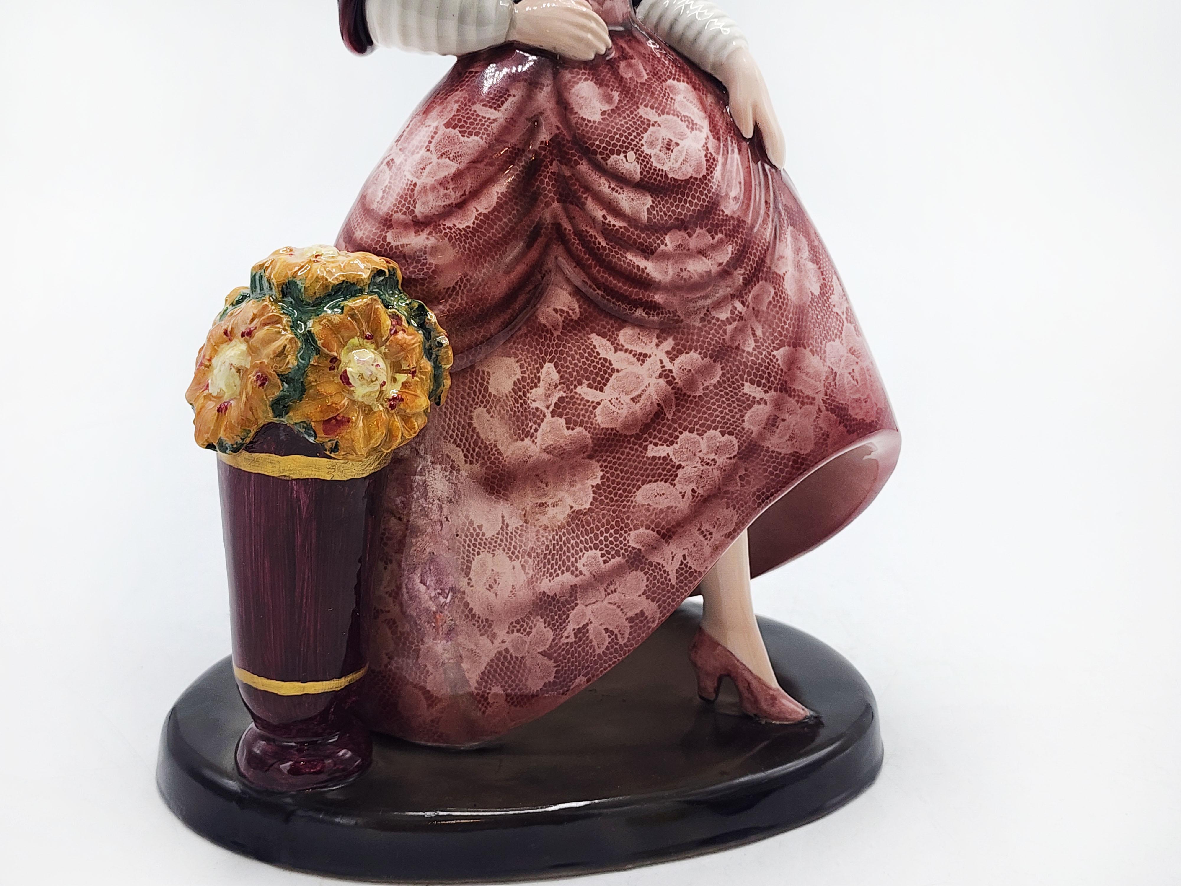 Un'imponente statuetta in porcellana Art Déco che cattura l'essenza della femminilità e dell'eleganza. La signora, con una postura aggraziata e un viso sereno, indossa un abito rosa pallido con intricati dettagli in pizzo, che vengono messi in