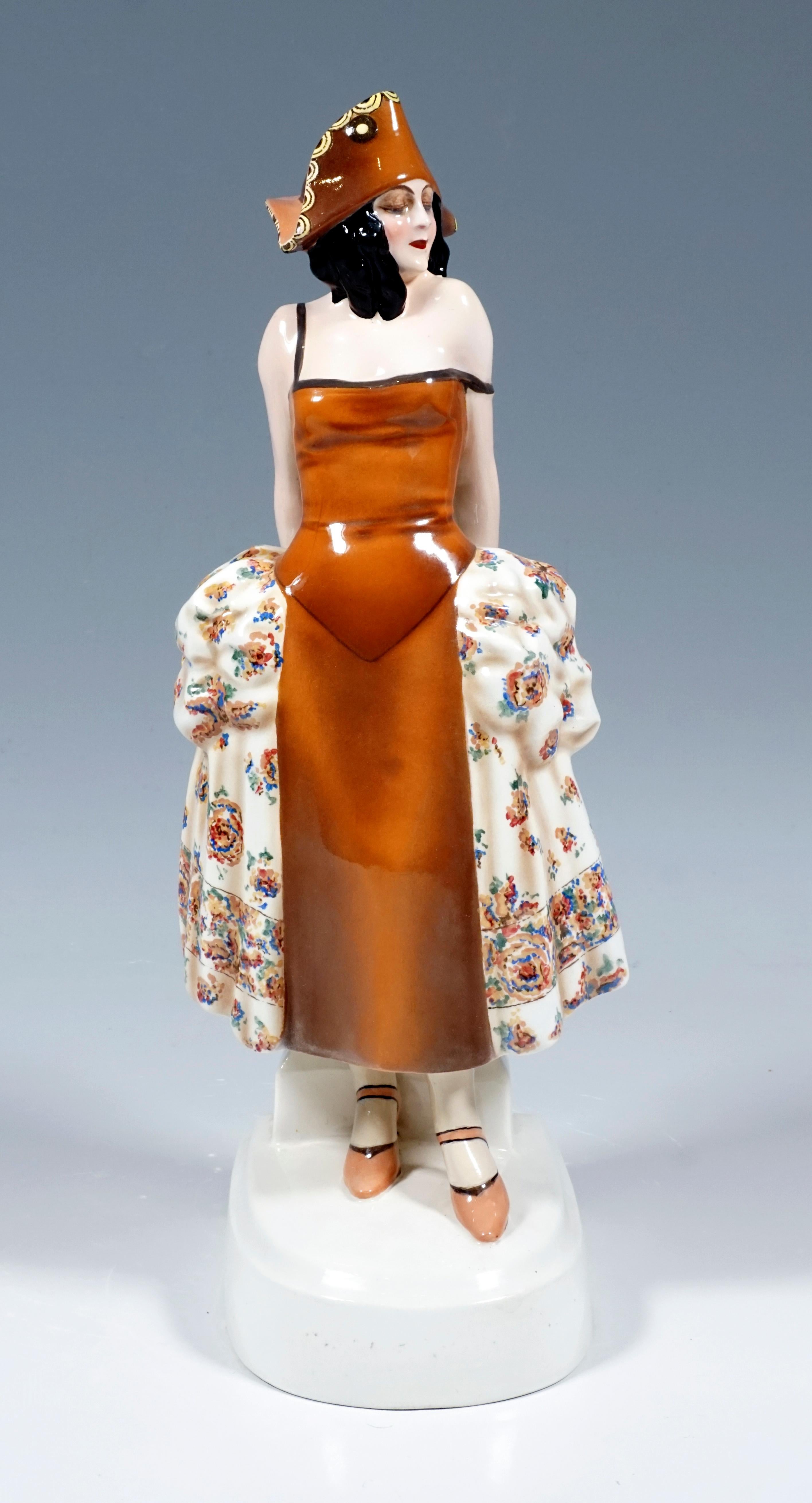 Rare Goldscheider Vienna Figurine of the 1920s:
Standing young lady in a brown carnival costume based on a rococo dress with a cream-colored skirt decorated with colored flowers and a brown tricorn, supporting herself with both arms on a column at