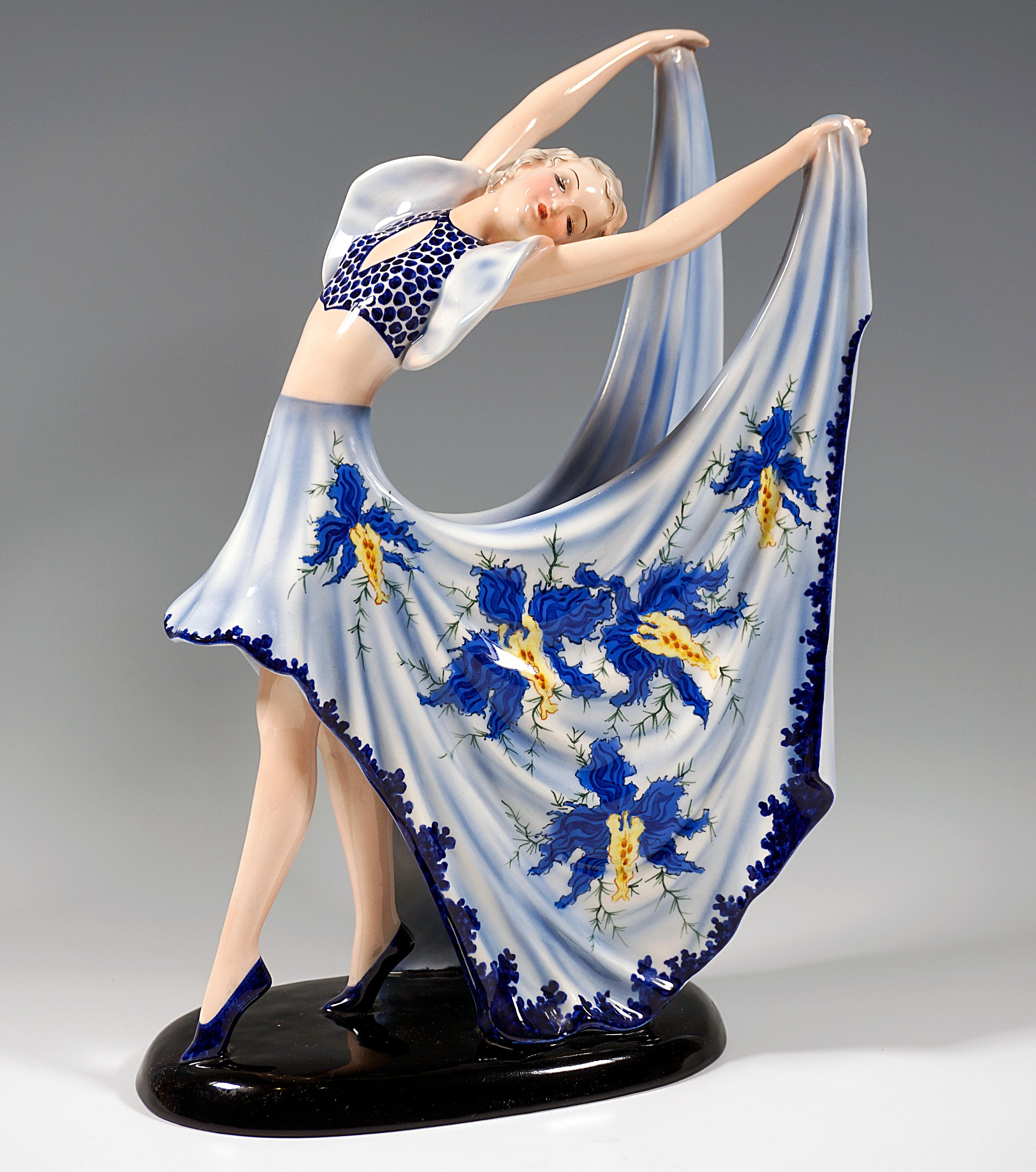 Gracefully posing dancer in blue costume: dark blue dotted bustier with light blue ruffled sleeves and short skirt in front with long, wide side parts with dark blue and yellow floral decoration slit at the back, holding these up backwards with arms