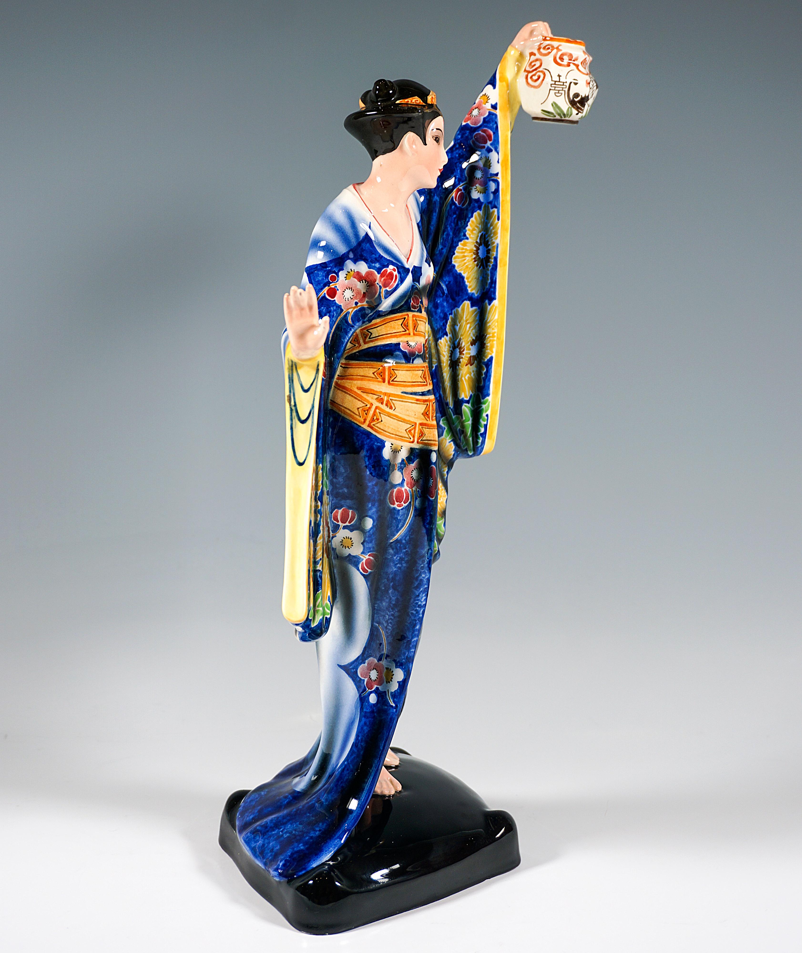 Rare and excellent Goldscheider art ceramic figure of the 1930's: 
Young standing Japanese woman in traditional kimono with wide sleeves and colored floral decoration on dark blue background, tiara in her upswept hair, looking to the left holding a