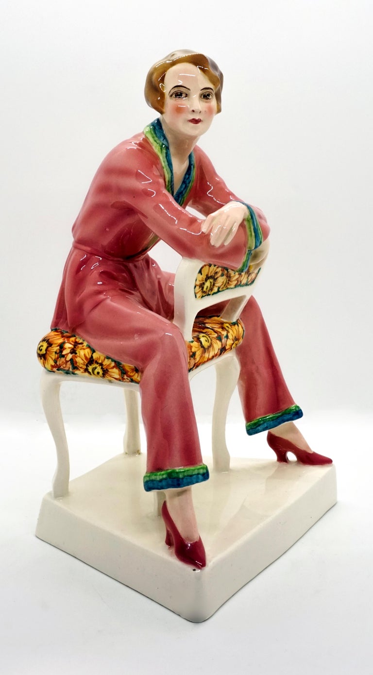 Very rare Goldscheider ceramic figurine.
The young lady with a brunette page-boy hair cut is wearing pink pajamas with blue-green hems and is sitting astride a white chair upholstered with a large floral pattern.
On a trapezoidal, cream-colored