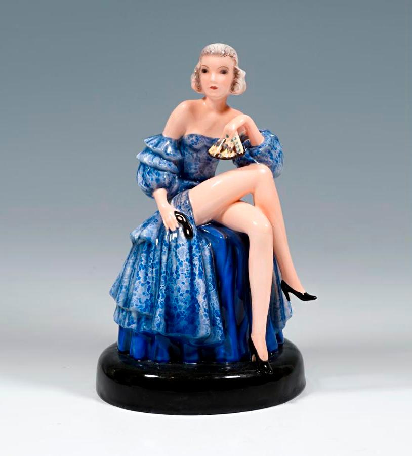 Rare Goldscheider Art Deco ceramic figurine:
Depiction of a seated dancer in a blue, strapless ball gown with lace flounces, the right leg folded over the left and the left arm on the knee, holding a ball mask and a fan in her hands.
The figure is