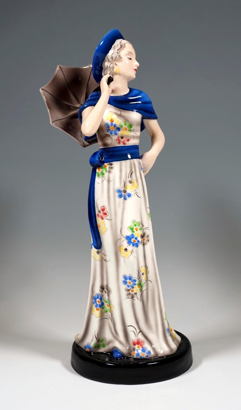 Very Rare Goldscheider Vienna Figurine of the 1930s:
Depiction of a standing, elegant lady in a floor-length, light, flowered dress with a blue shawl collar, belt strap and hat, resting her left hand on her hip, laying a small gray parasol on her