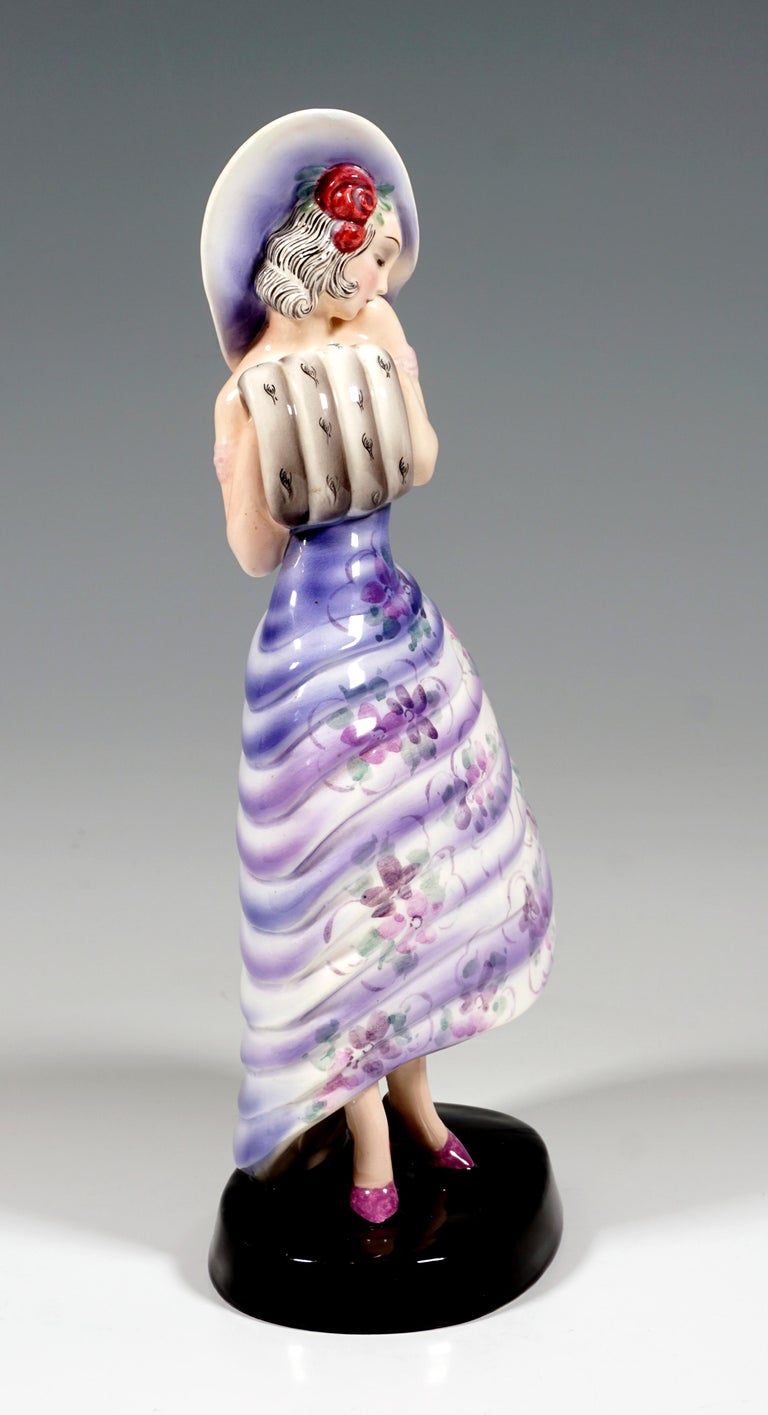 Rare Goldscheider Art Deco ceramic Figurine:
Representation of an elegant, standing lady in a dress with a wide flounced skirt with a stylized flower pattern in violet and rose tones, red roses adorn her shoulder-length hair, a wide-brimmed hat