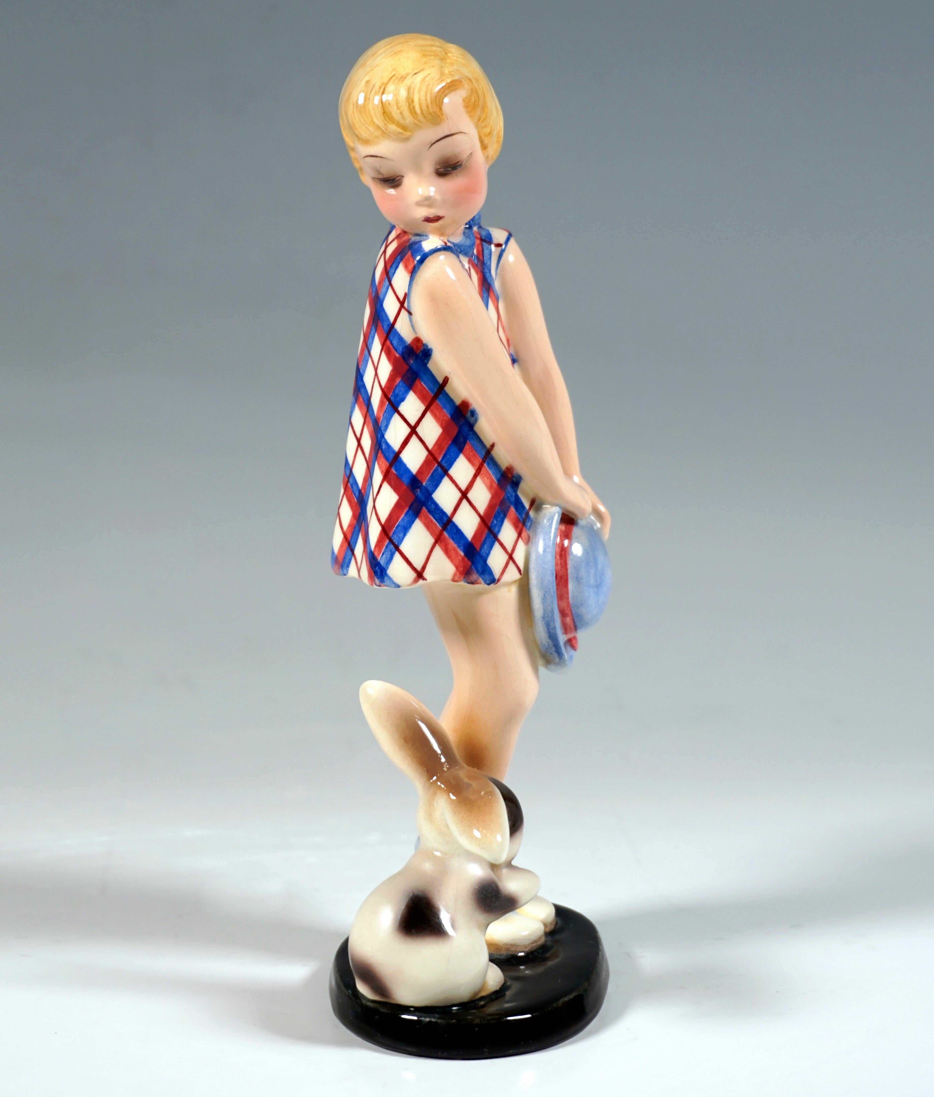 Little blond girl in red and blue plaid dress, holding a light blue hat in both hands in front of her, looking over her right shoulder down at a small rabbit beside her.
On a black, oval flat base.

Designer:
Claire/Klára Herczeg/Weiss (1906 -