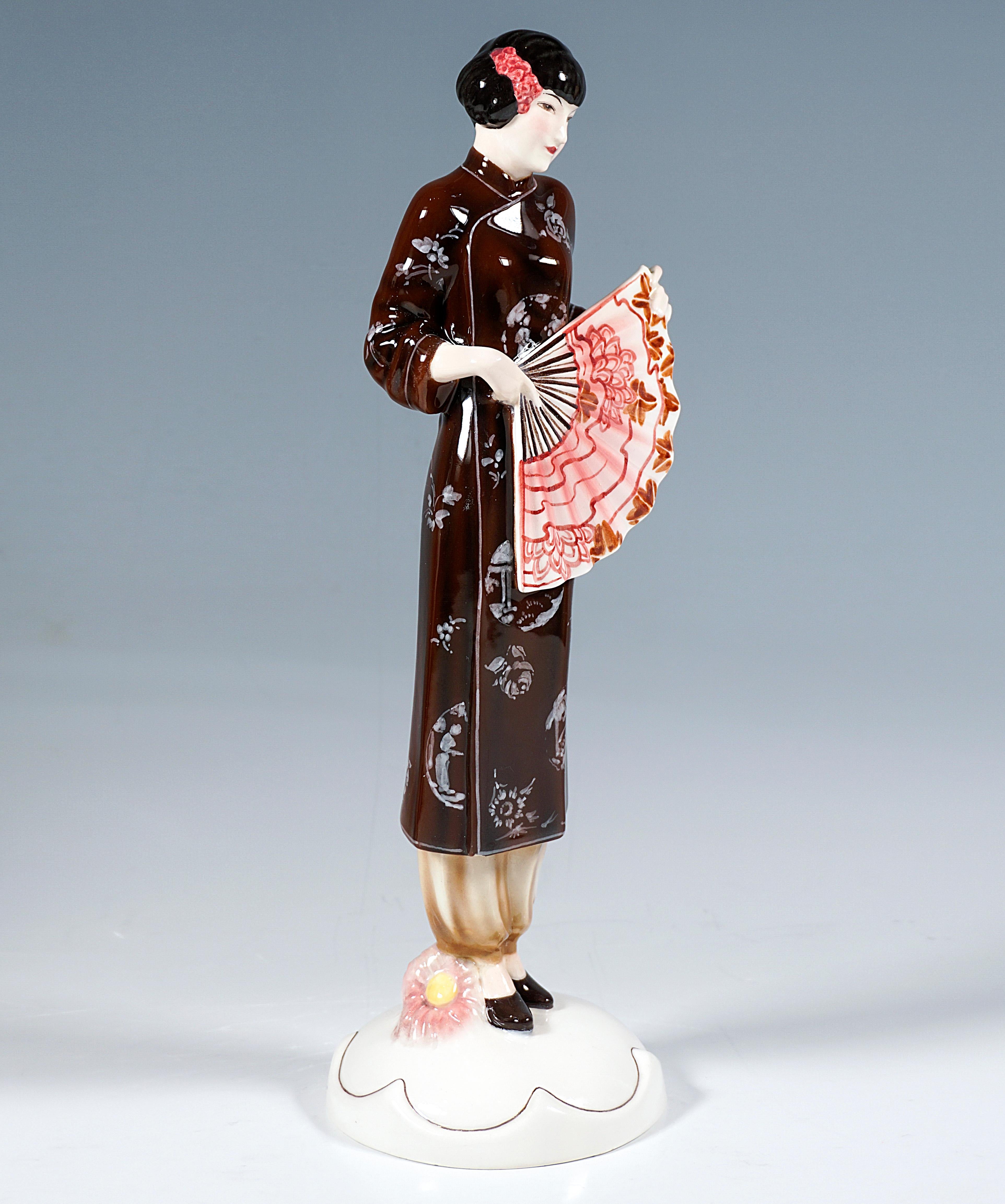 Very rare Goldscheider Vienna Ceramic Figurine of the 1930s:
Representation of a young lady in a brown, Japanese silk coat over light, baggy trousers, hair pinned up with floral decoration, standing upright and holding a large fan in both hands in