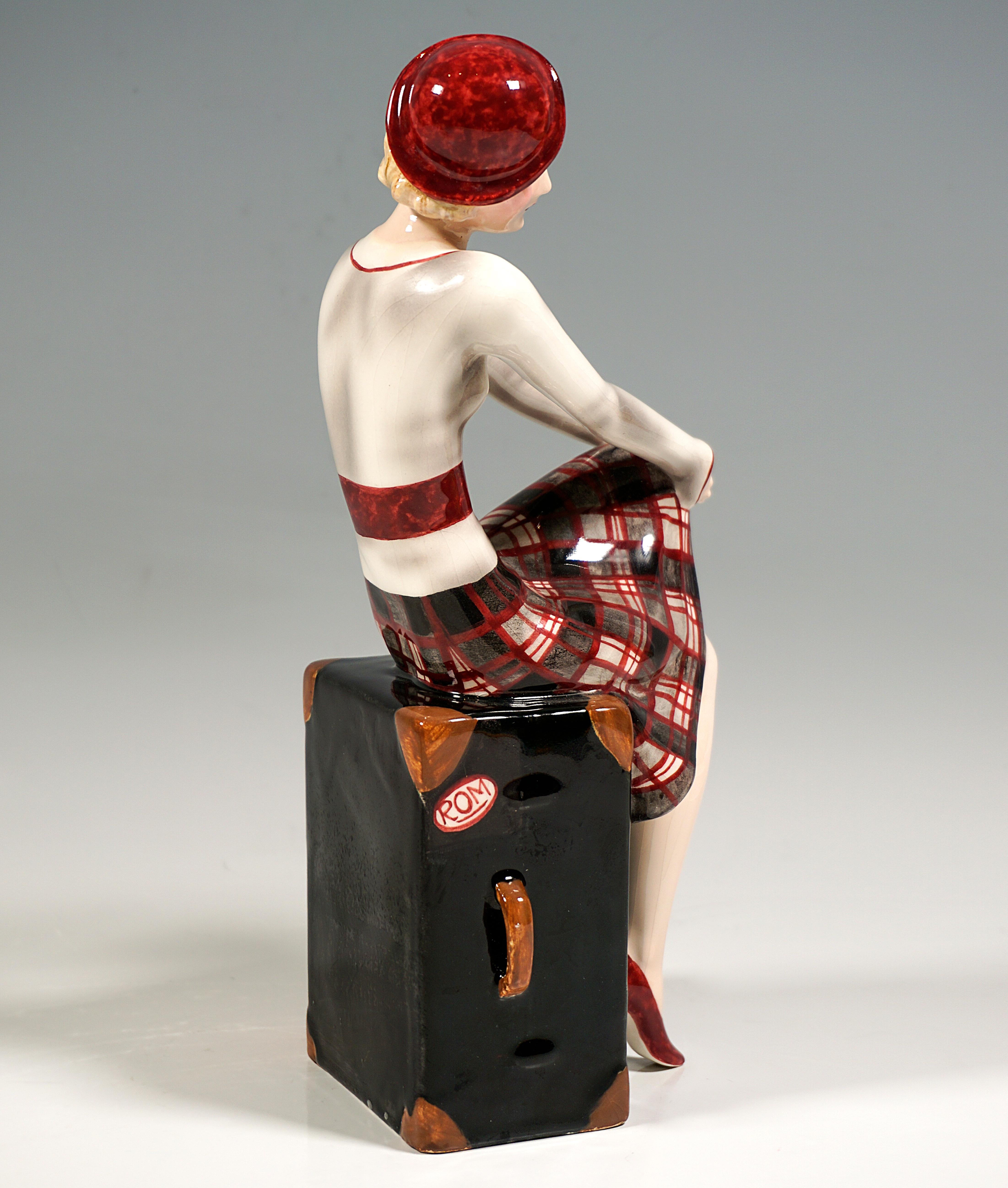 Very Rare Goldscheider Art Déco Ceramic Figurine Of The 1930s:
Blond young lady with a small red hat, in a light gray vest over a white blouse and in a gray, white and red checked skirt, sitting on a large black suitcase, pulling up her left leg