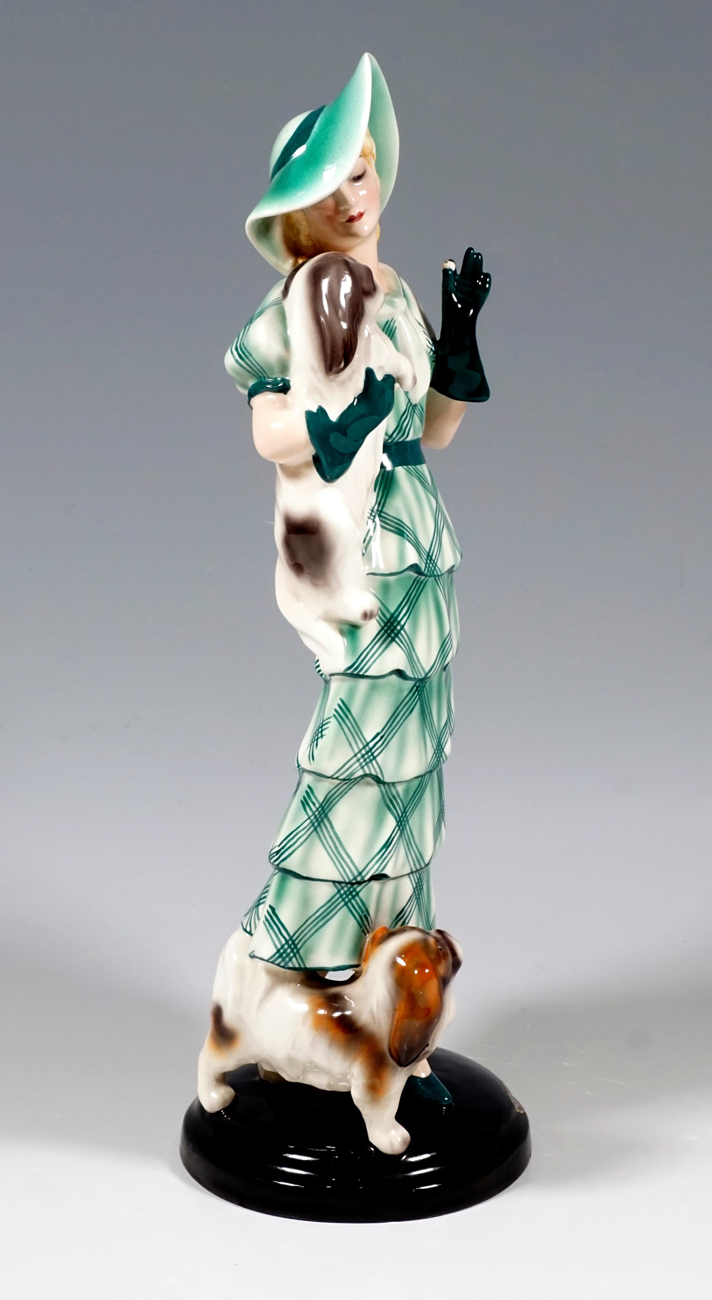 Very rare Goldscheider Vienna figurine of the 1930s:
Representation of a standing elegant lady in a long, beige, green checked dress with stepped flounces, matching hat and green gloves, holding a Pekingese in her right arm and offering him a treat