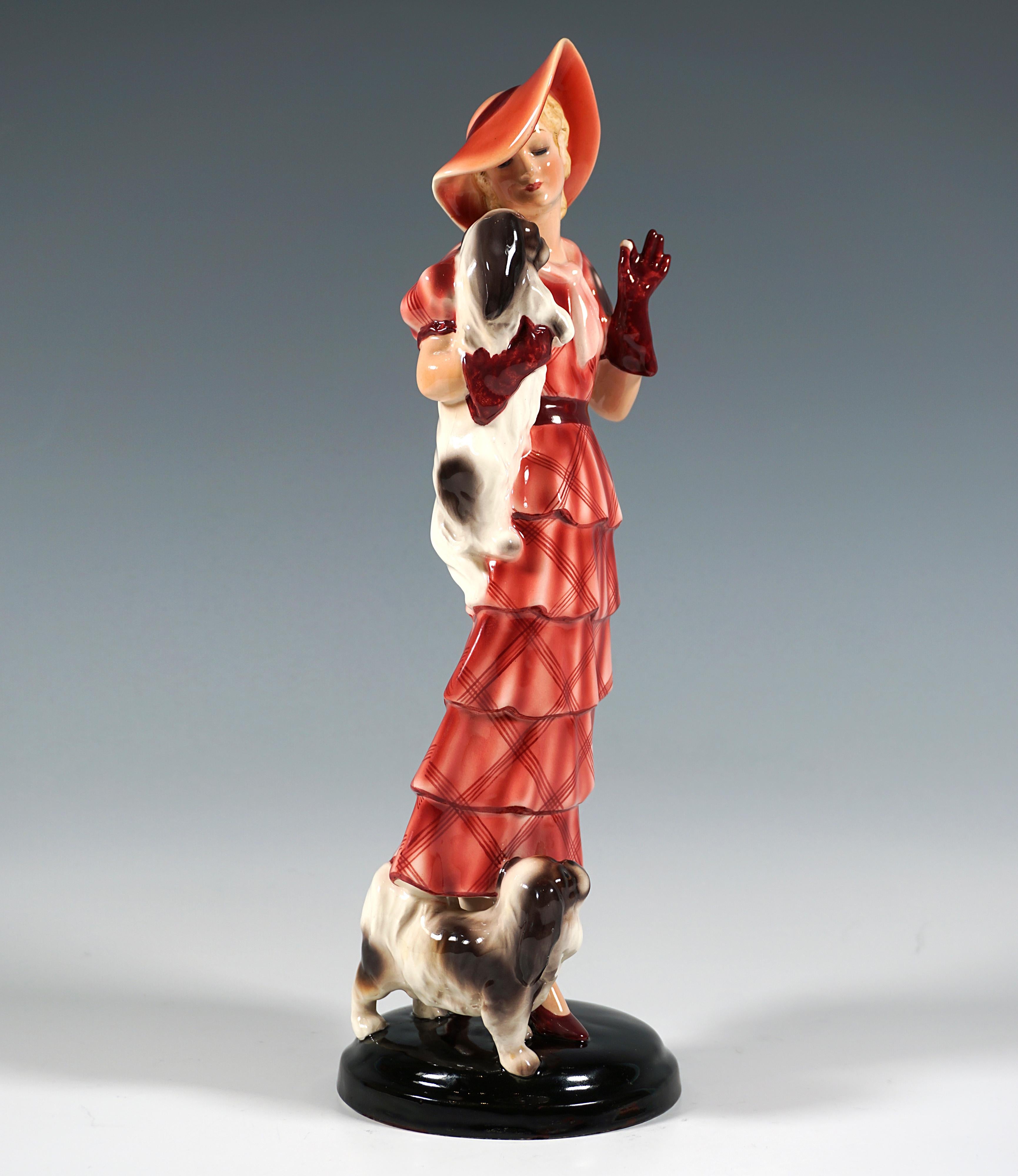 Very Rare Goldscheider Vienna Figurine of the 1930s:
Depiction of a standing, elegant lady in a long, red, checkered dress with tiered flounces, matching hat and red gloves, holding a Pekingese in her right arm and offering it a treat with her left
