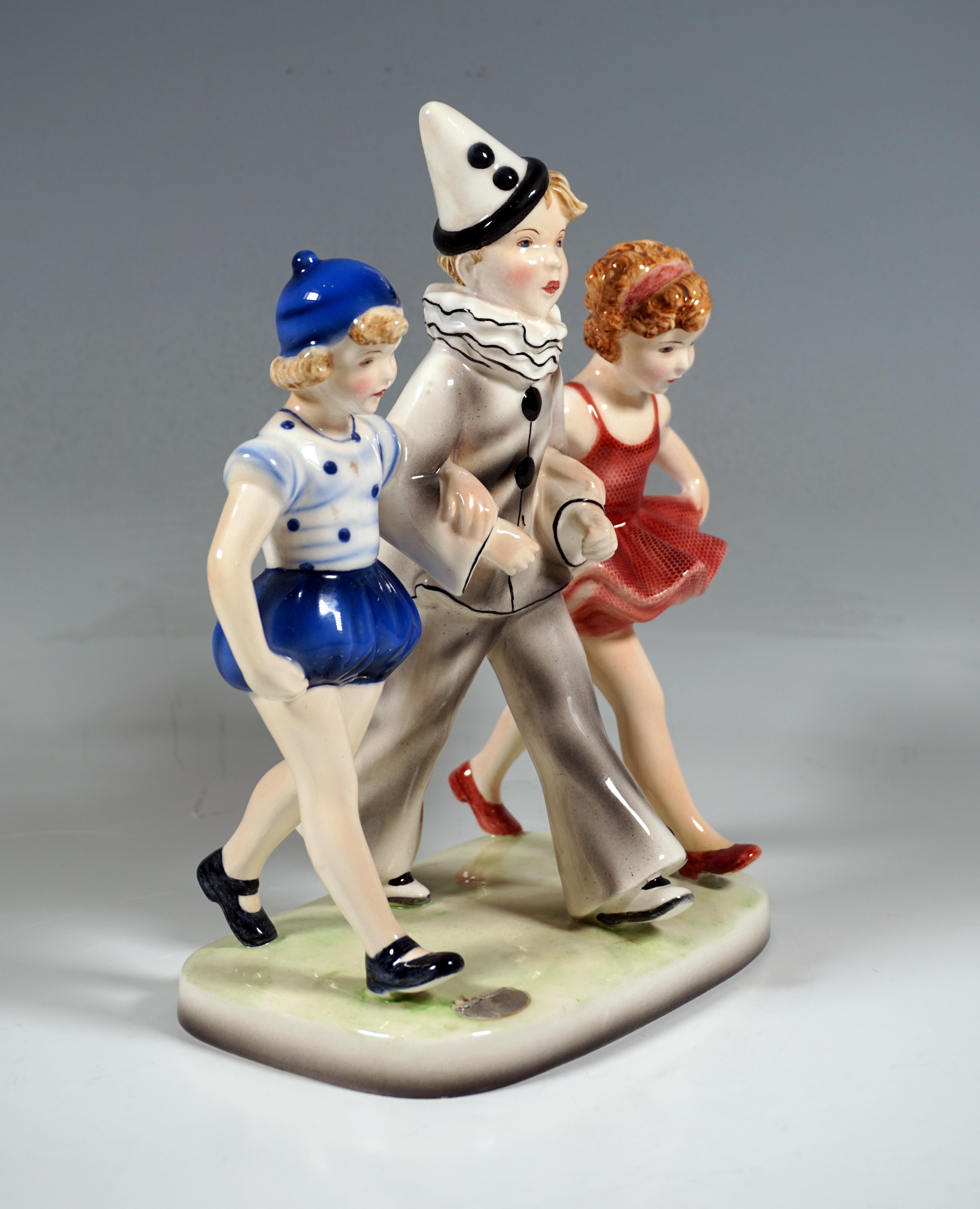 Three children walking side by side: in the middle a blond boy dressed as Pierrot with a pointed hat, arm in arm with two girls, one in a red tutu dress, the other in a blue dotted camisole with matching short harem pants and a bonnet.
On a