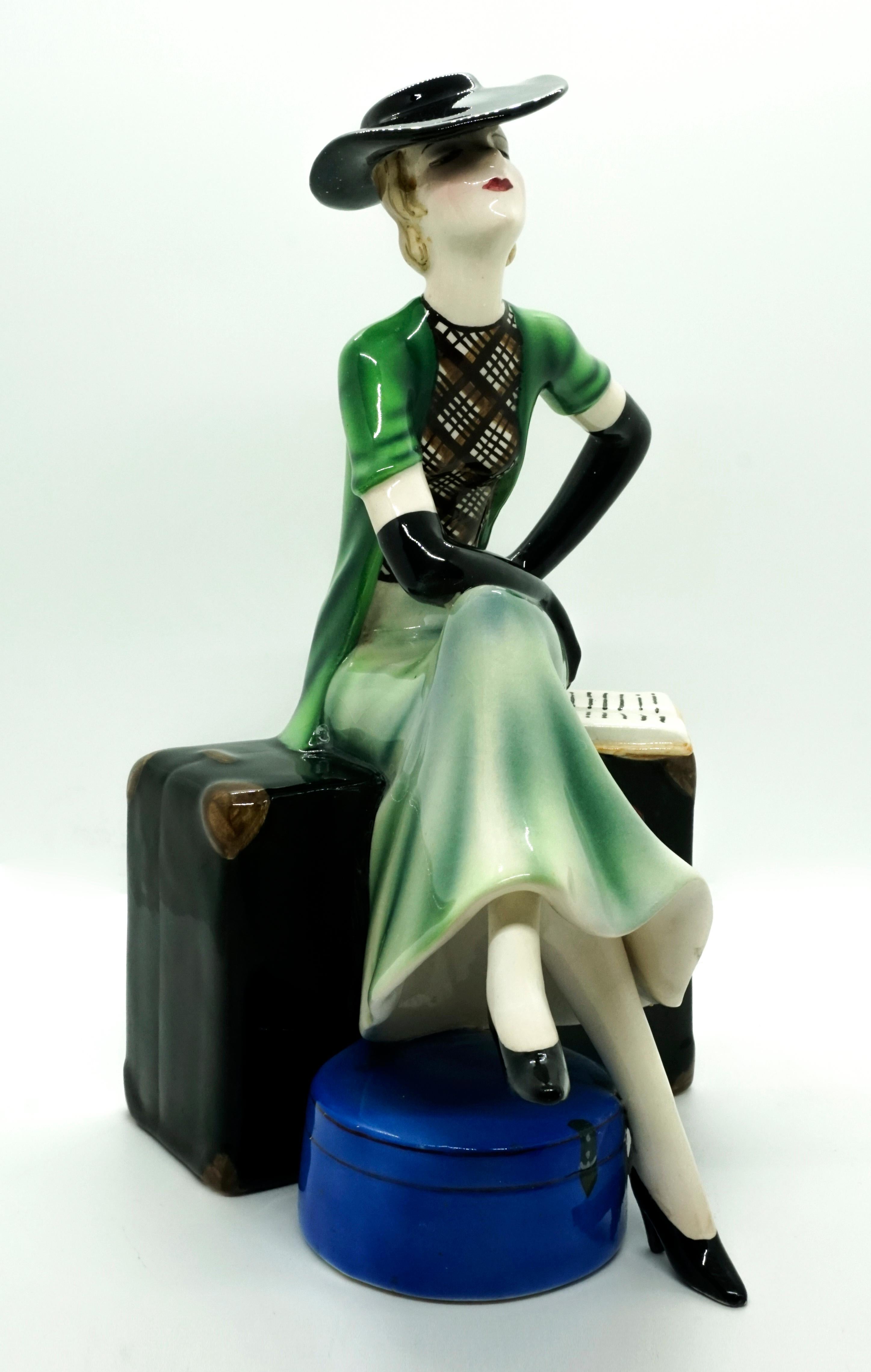 Rare Goldscheider Art Deco ceramic figurine.
A young lady with chin-length blond hair and an elegant black hat, a long green skirt, a checked blouse, over it a short-sleeved green vest and long black gloves is sitting, her legs crossed and her left