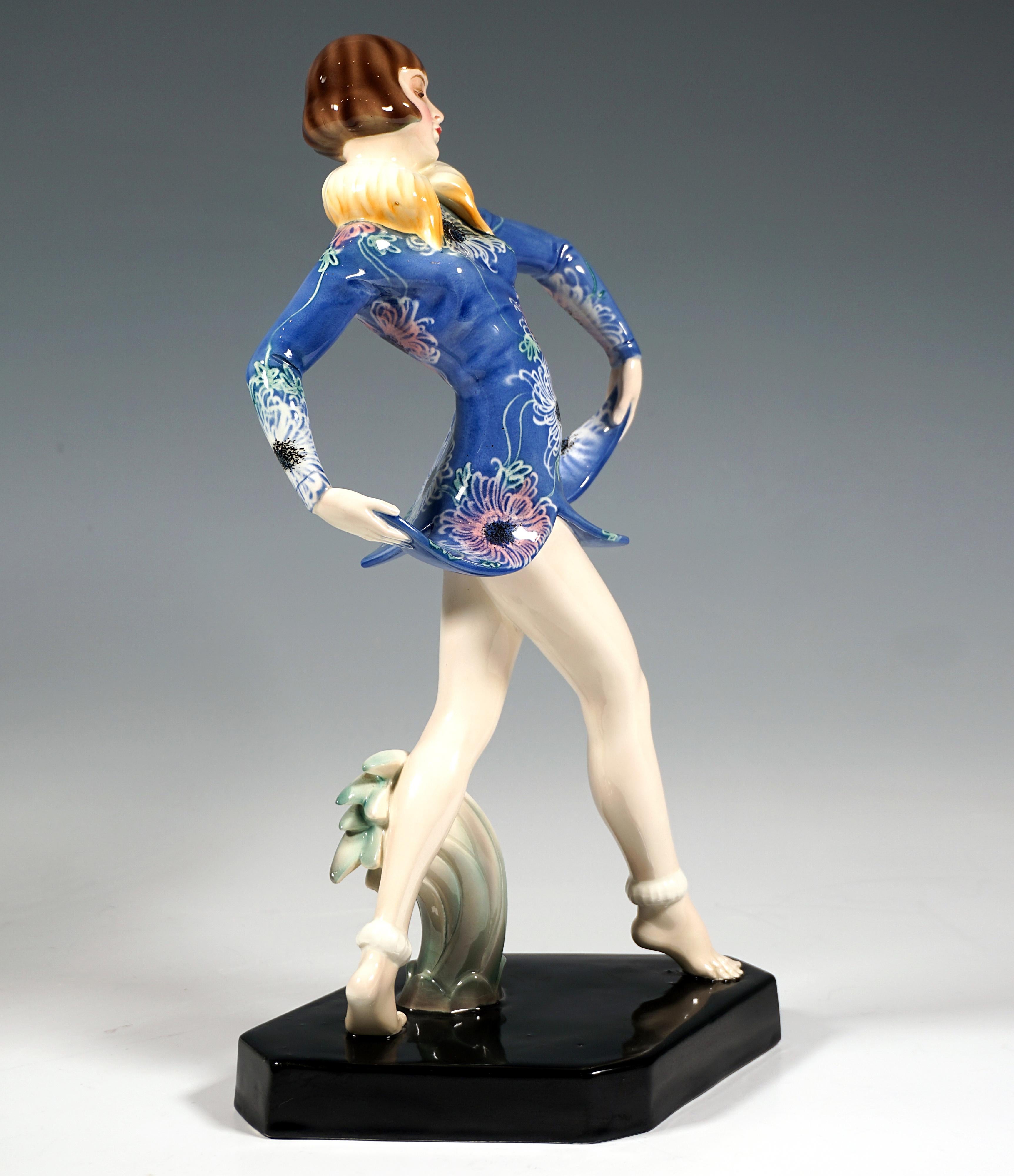 Joven bailarina de revista con peinado de paje, dando un paso adelante mientras levanta el dobladillo en forma de campana de su vestido azul corto con cuello de piel y colorida decoración floral estilizada con ambas manos hacia los lados, adelante y