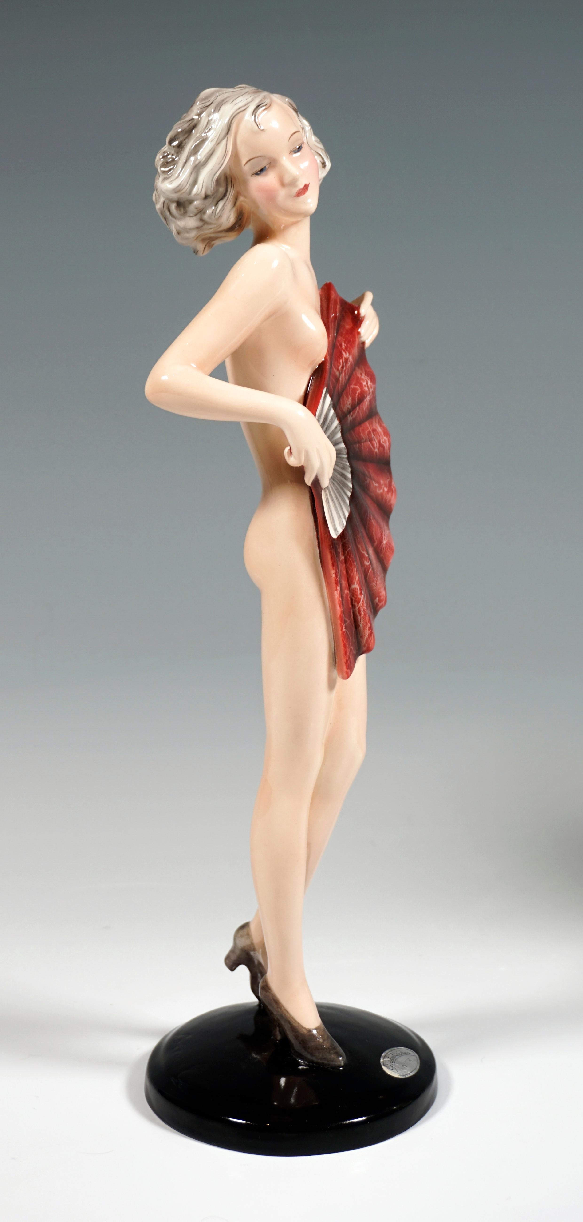 Very rare Goldscheider ceramic figurine of the 1930s:
The standing young pretty lady with curly, chin-length hair only wears high heels and covers her nakedness with a large, red fan which she holds in front of her with both hands.
On a black,
