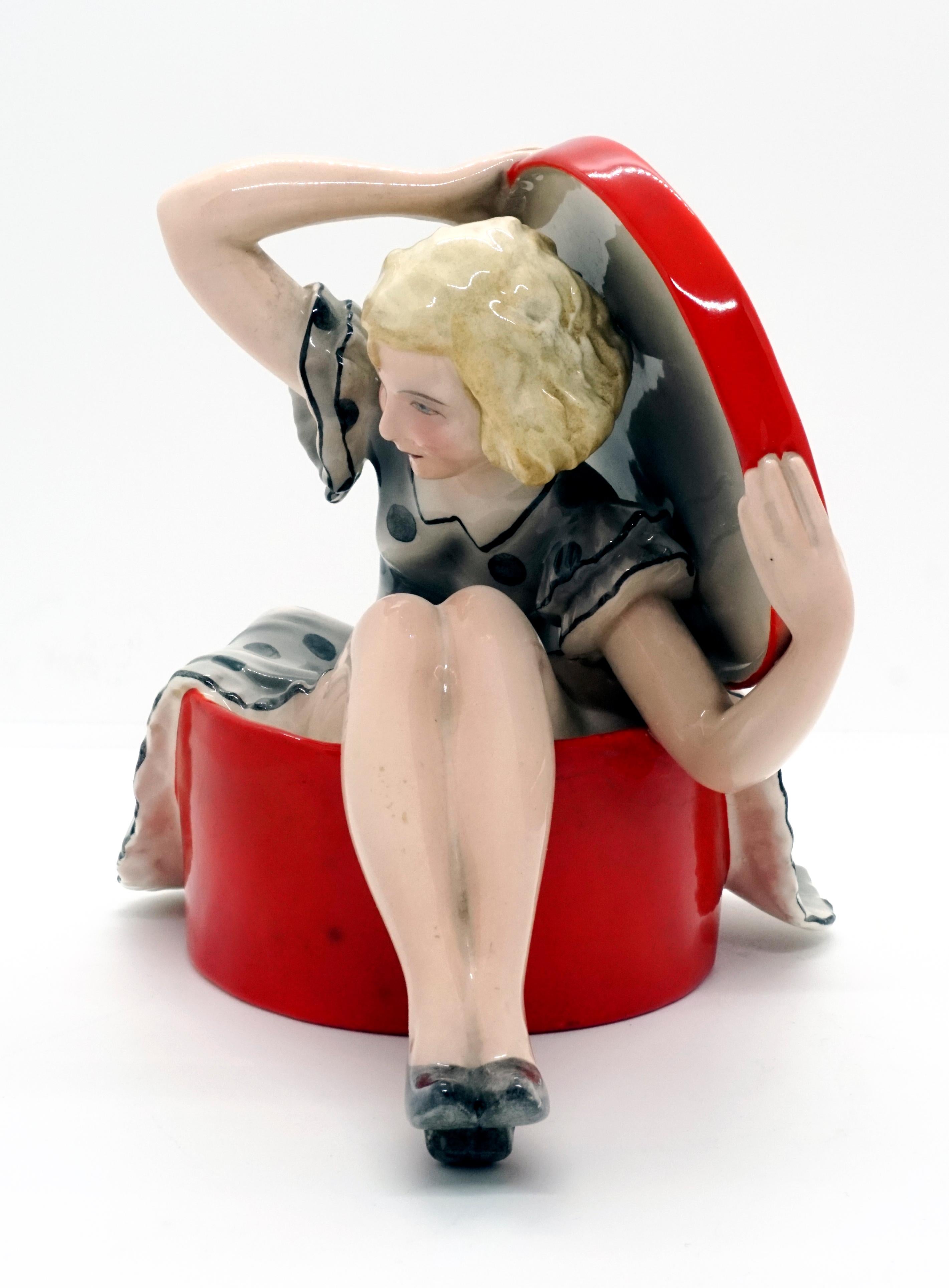 Very rare Goldscheider Art Deco ceramic figurine.
A young lady with blond curly hair and a light gray, spotted summer dress is sitting in a large, red hat box, leaning slightly forward, holding the lid up behind her with both hands.

Designed by