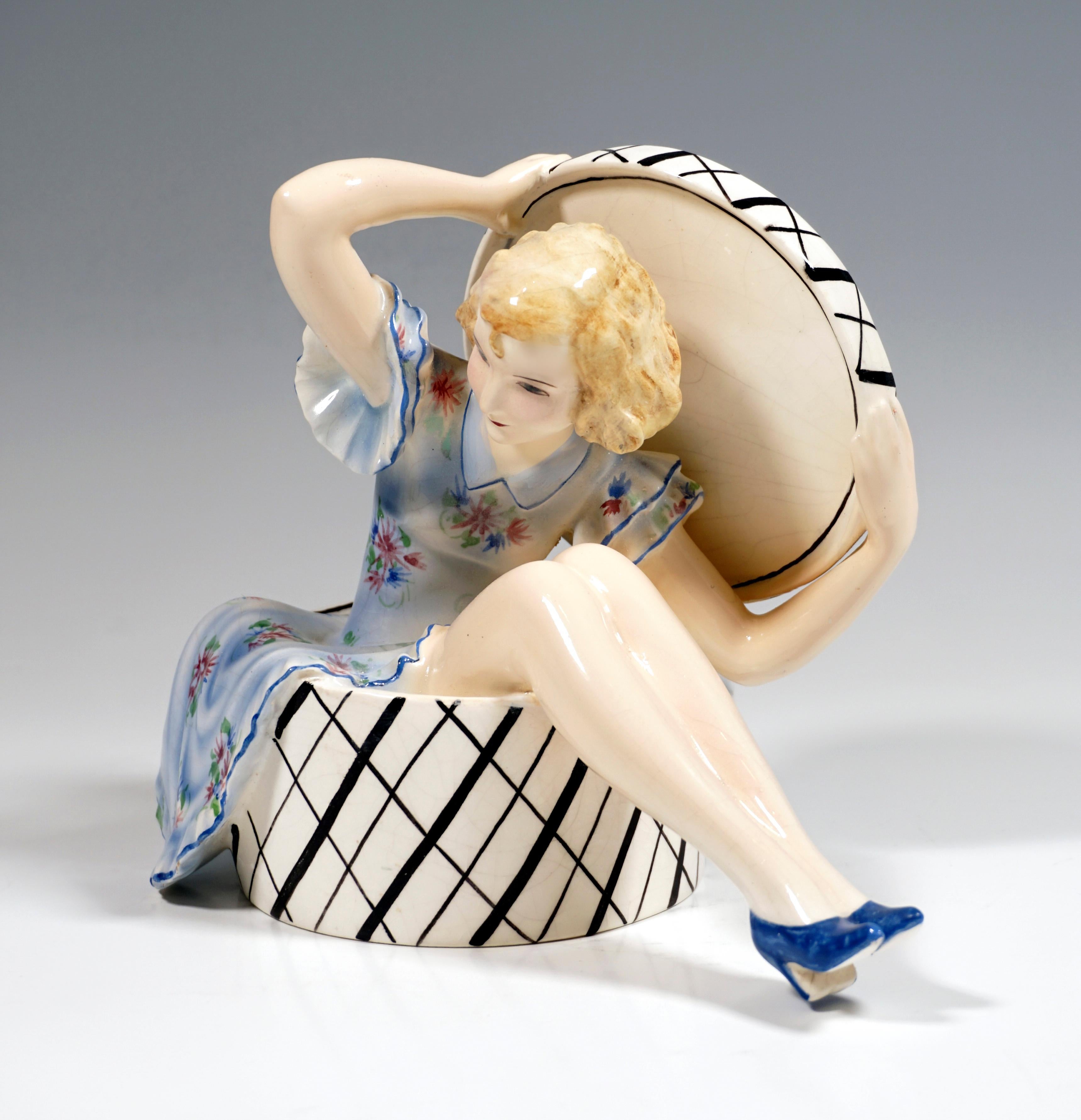 Very Rare Goldscheider Art Déco Ceramic Figurine.
A young lady with blond curly hair and a light blue, floral summer dress sitting in a large, white and black checkered hat box, leaning slightly forward, holding the lid up behind her with both