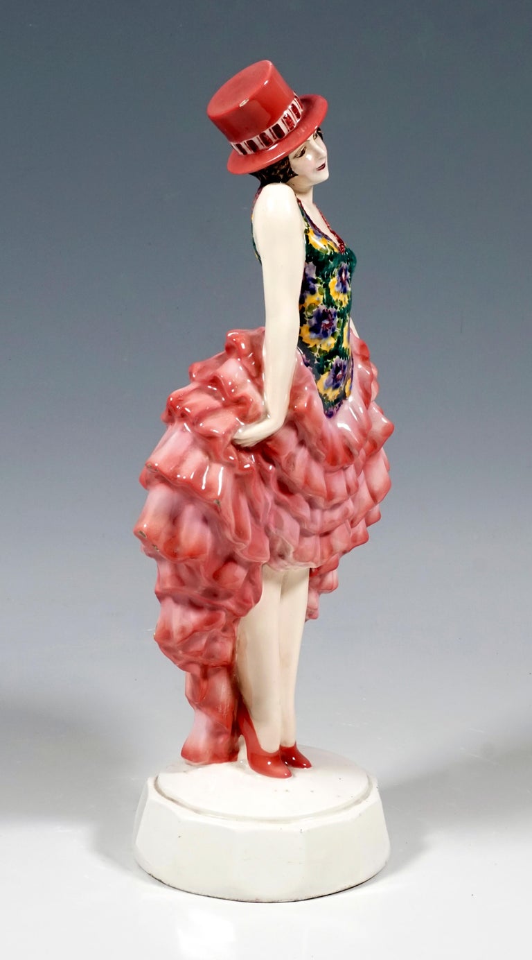 Rare goldscheider figure of the 1920s:
The standing dancer wears a pink cylinder hat on her short brown locks. The flower-patterned top of her costume is tight-fitting and sleeveless. Tilting her head slightly to the right, she pulls up her