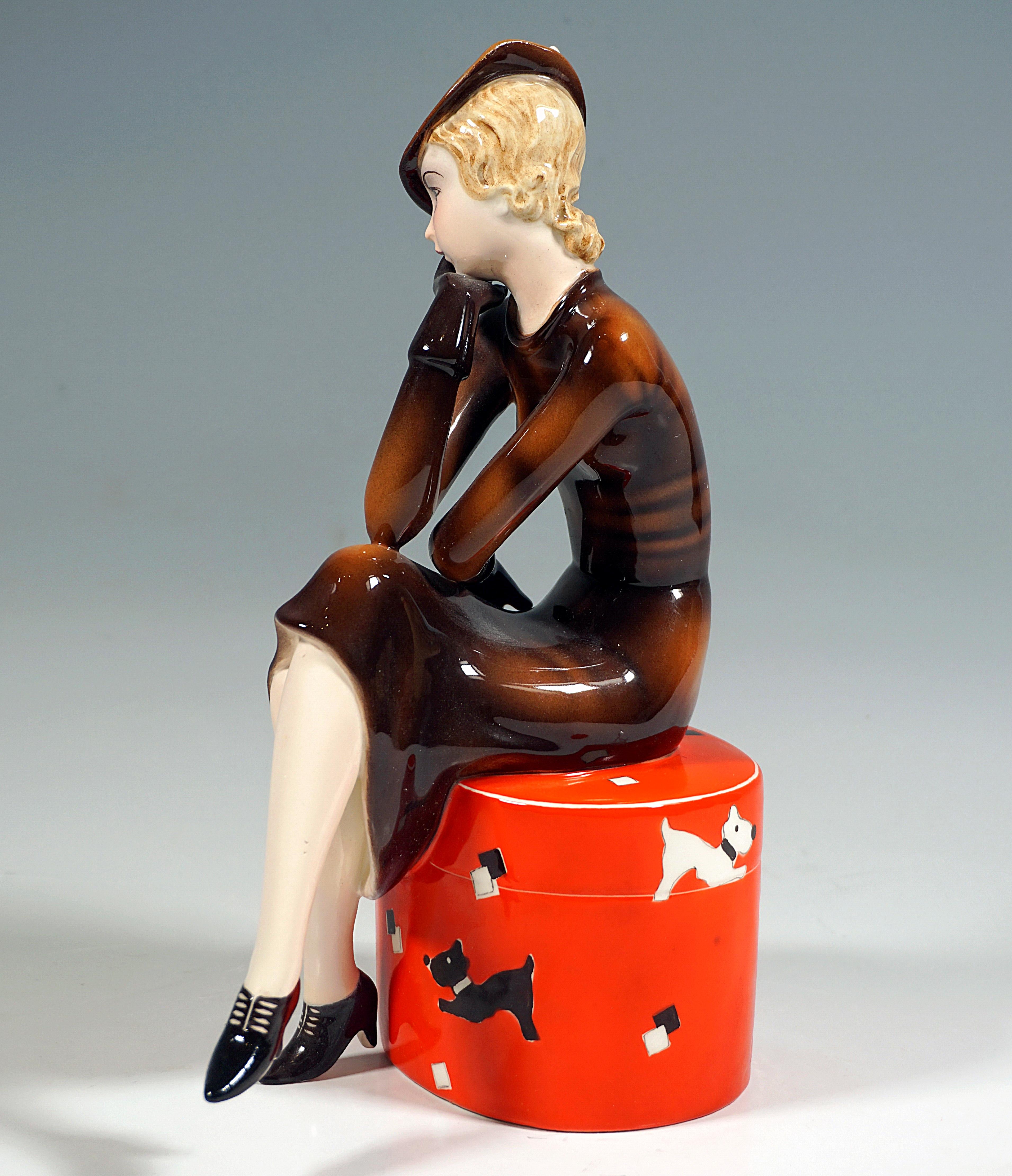 Very rare Goldscheider Art Deco ceramic figurine:
A young lady with blond curly hair in an elegant, brown suit with a matching hat and gloves sitting on a large, red, round box with black and white dog decorations, with crossed legs, her head