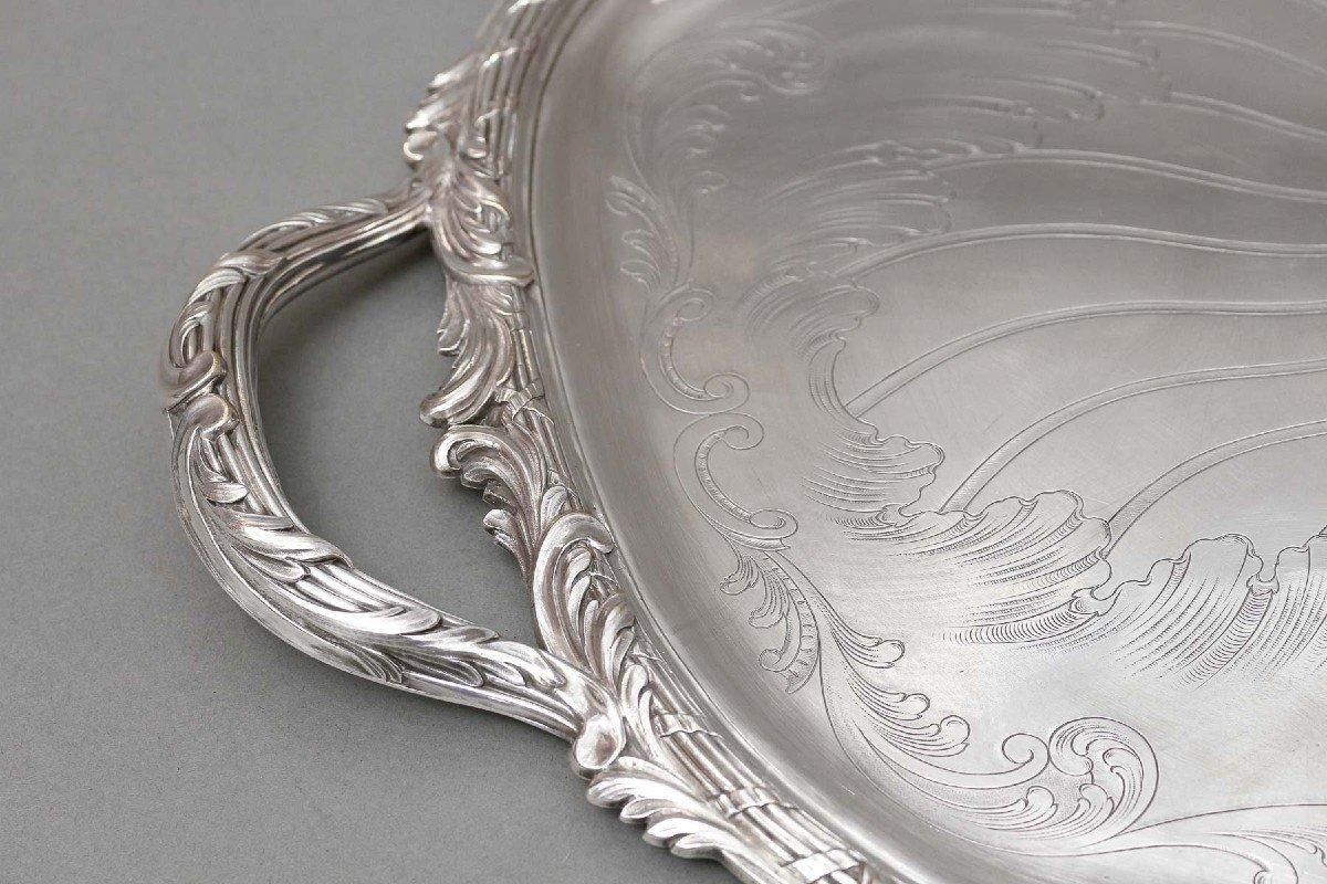 Oval tray in silver-plated metal, with an animated shape, interior chiseled with a radiant decoration surrounding a cartouche, stylized arabesques, and flanked by two side handles. The edge with crossed nets and ribbons is decorated with foliage.