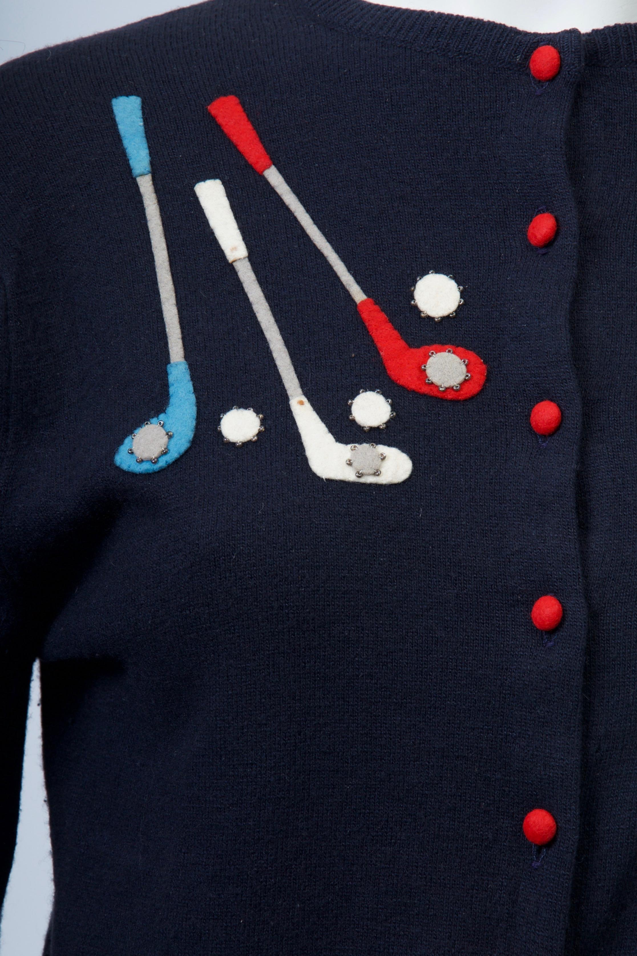 Decorated cardigans are some of the most iconic vintage items from the 1950s and '60s. This example is fashioned of navy cashmere and features felt appliqués in white, blue and red of golf clubs, club covers, and a golf bag. Tiny red buttons stand