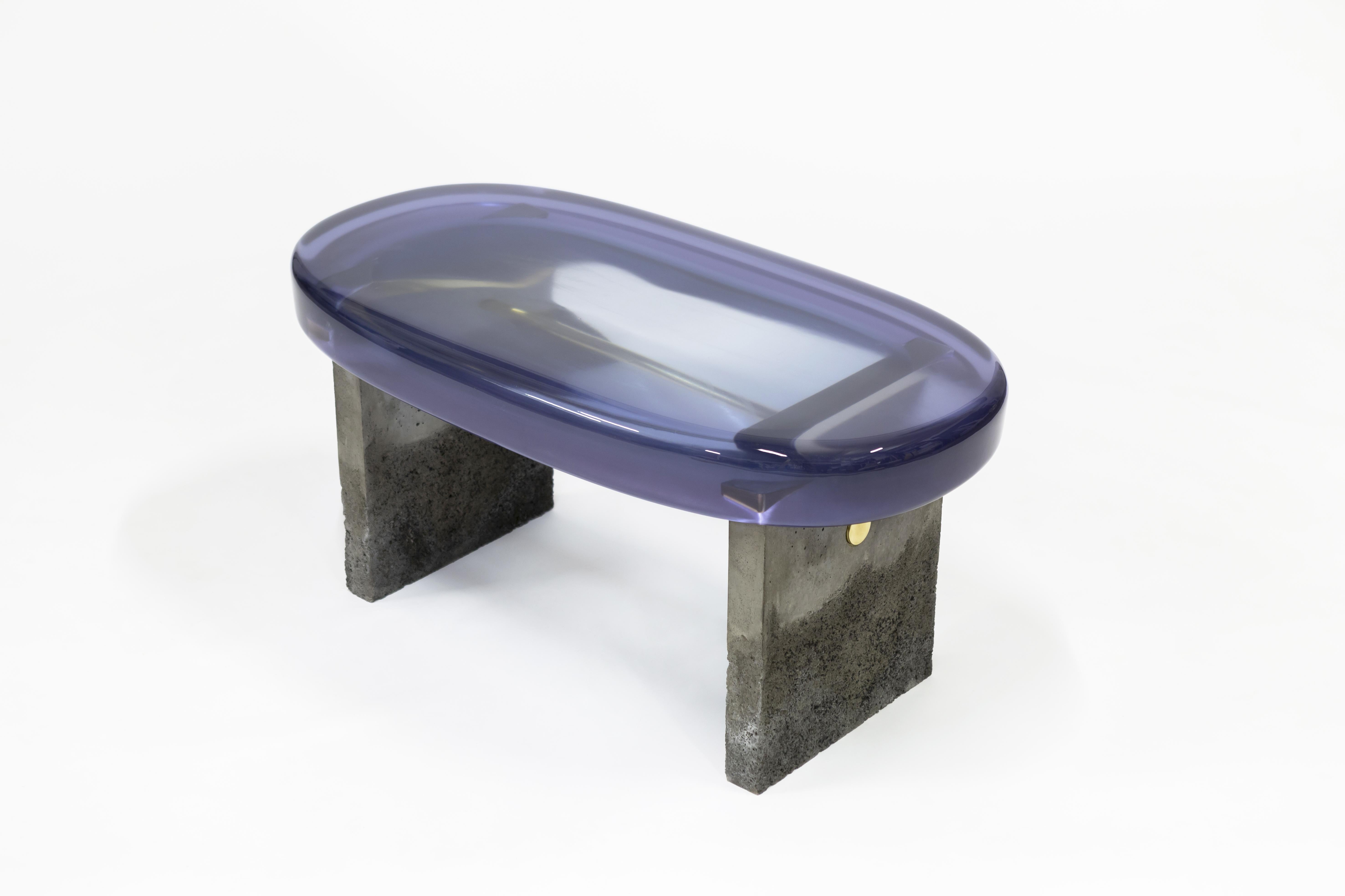 D'une composition formelle stylisée, la banquette Golia présente une épaisse assise ovale en résine époxy soutenue par deux pieds carrés en béton, maintenus par une barre en laiton. La résine transparente révèle les liens métalliques brillants