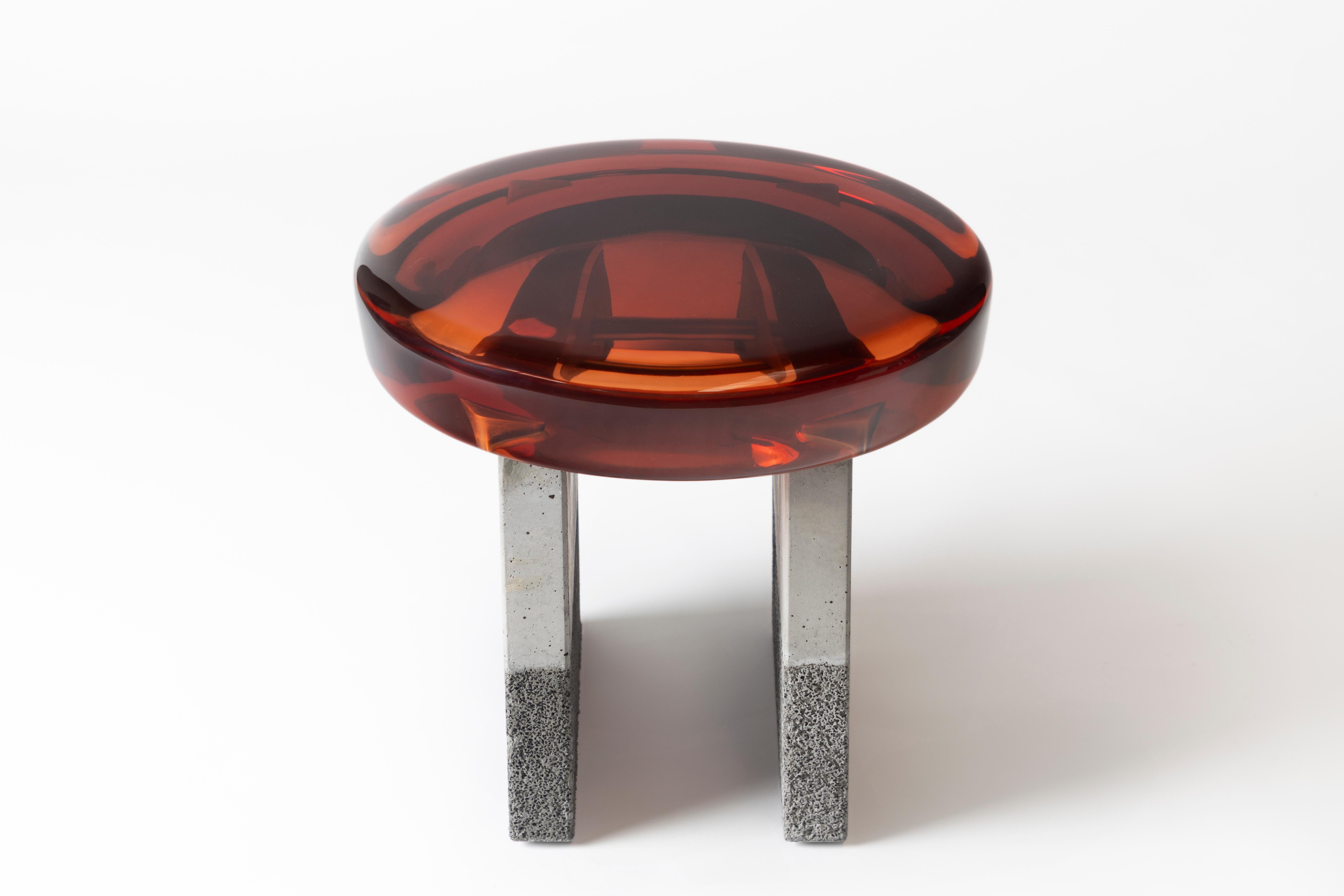 D'une composition formelle stylisée, le tabouret Golia présente une épaisse assise ronde en résine époxy reposant sur deux pieds carrés en béton. La résine transparente révèle les liens métalliques brillants situés en dessous, créant une vue à