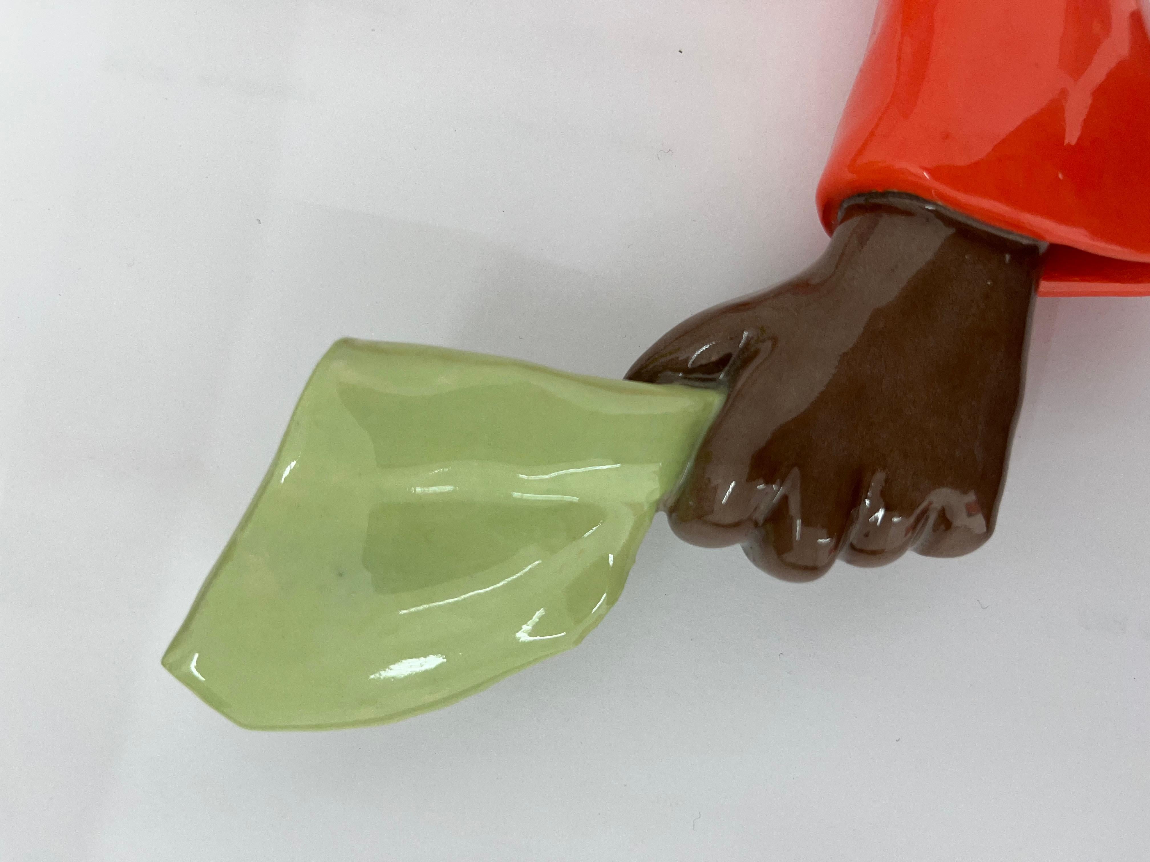 This glazed ceramic sculpture presents an elongated orange arm that extends outward, ending in a brown hand firmly gripping a green machete. The exaggerated proportions distort the natural scale of the human body, emphasizing the gesture and the