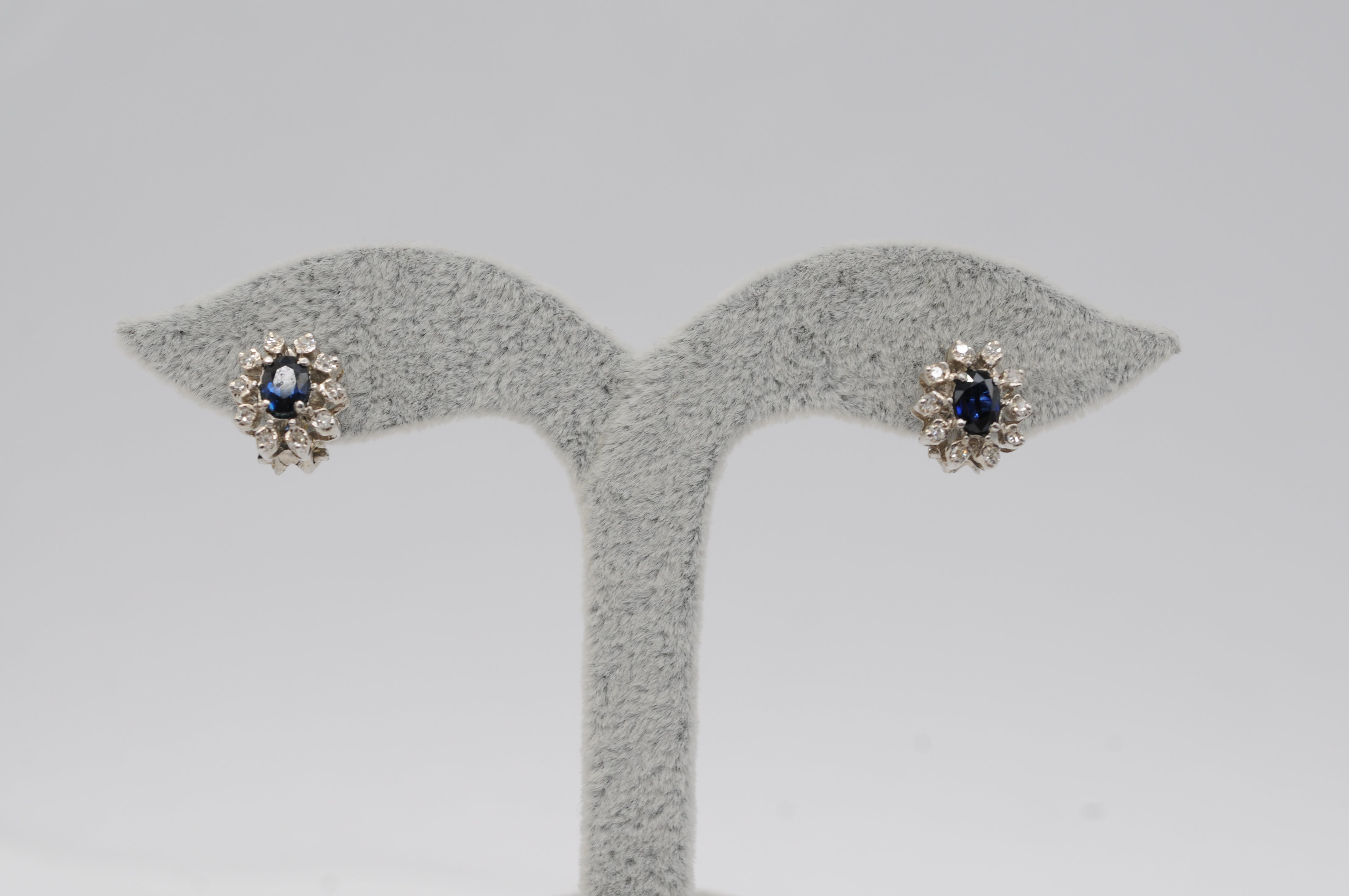 Lassen Sie sich von der schieren Schönheit dieses atemberaubenden Ohrringpaars verzaubern. Jeder Ohrring ist aus exquisitem 14-karätigem Weißgold gefertigt und zeigt einen bezaubernden Saphir im Ovalschliff, der von insgesamt 10 Diamanten im