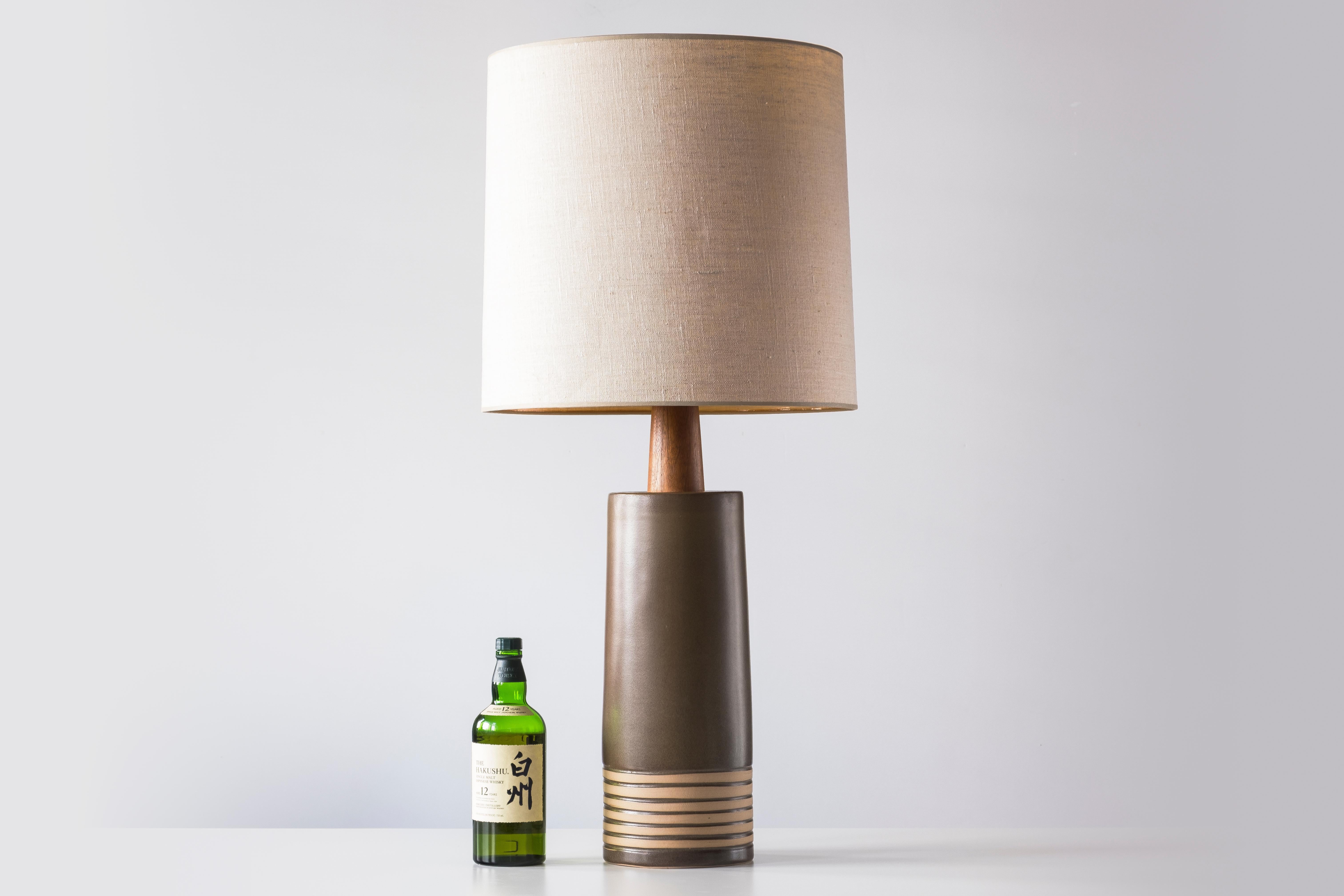 WHAT IS IT?
—
This signed Martz Model 223 lamp comes in matte olive glaze with six stripes towards the base revealing unglazed clay. The tall ceramic body gives way to a thick, tapered walnut neck and the whole ensemble is capped off with an