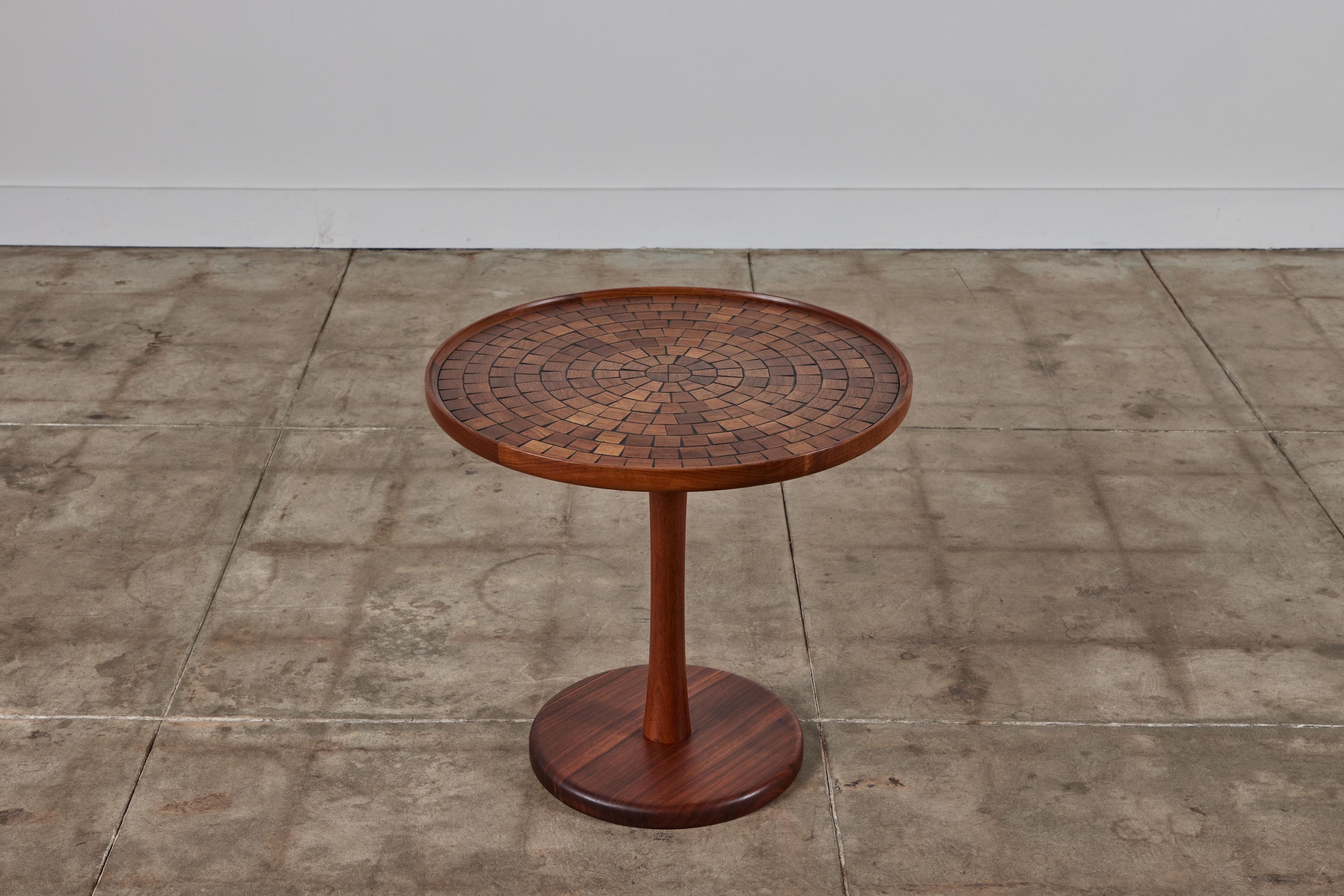 Round oak side table by Gordon & Jane Martz. The table top is inlaid with square and triangular oak tiles in a geometric pattern. The tiles feature varying wood grains and brown tones . The frame, base and pedestal of the table are all solid