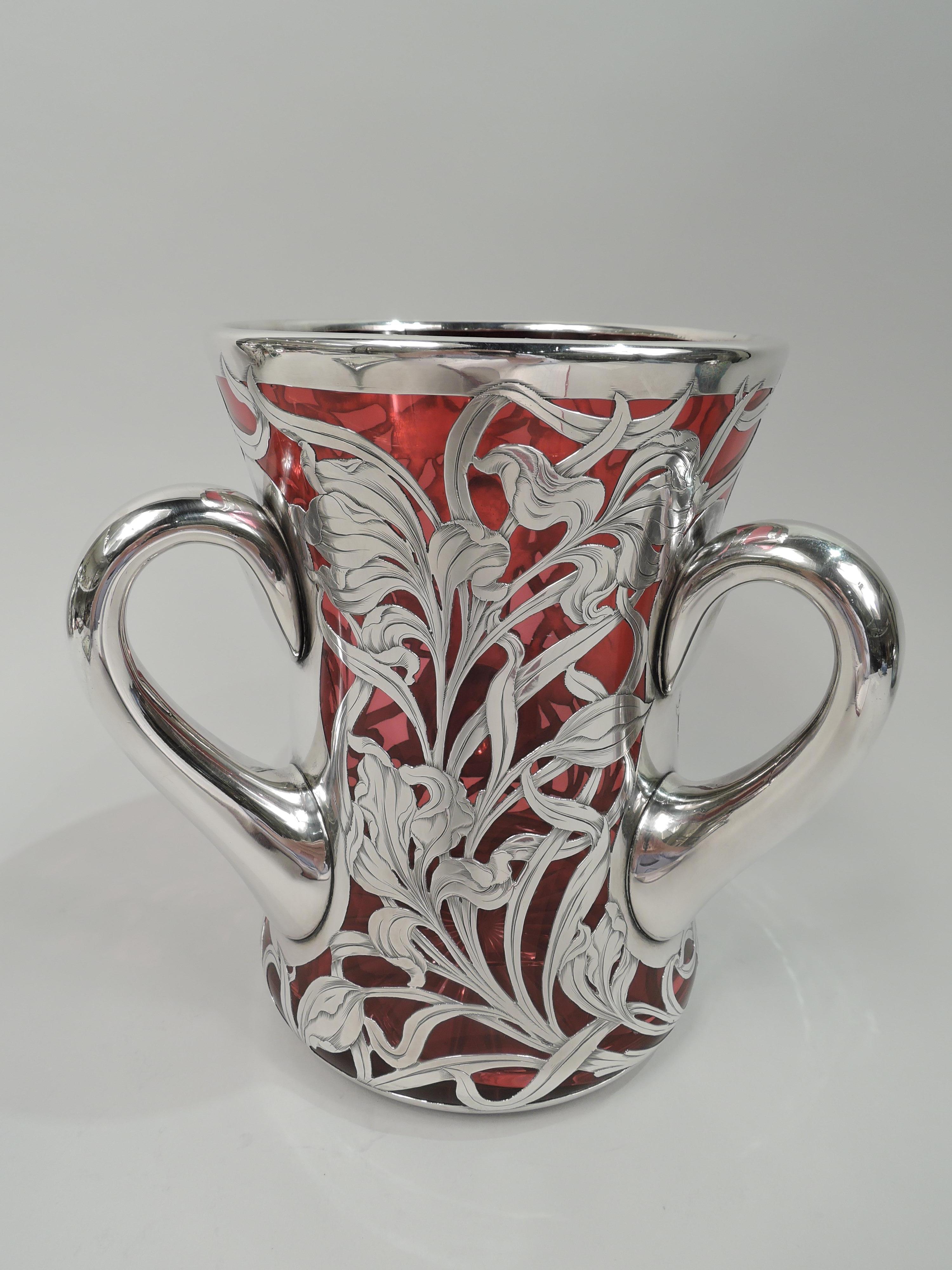 Gorgeous turn-of-the-century American Art Nouveau glass loving cup with engraved silver overlay. Waisted cylinder with looping c-scroll silver handles. Clear-cut star on underside. Overlay in form of wildly windswept and entwined iris flowers and