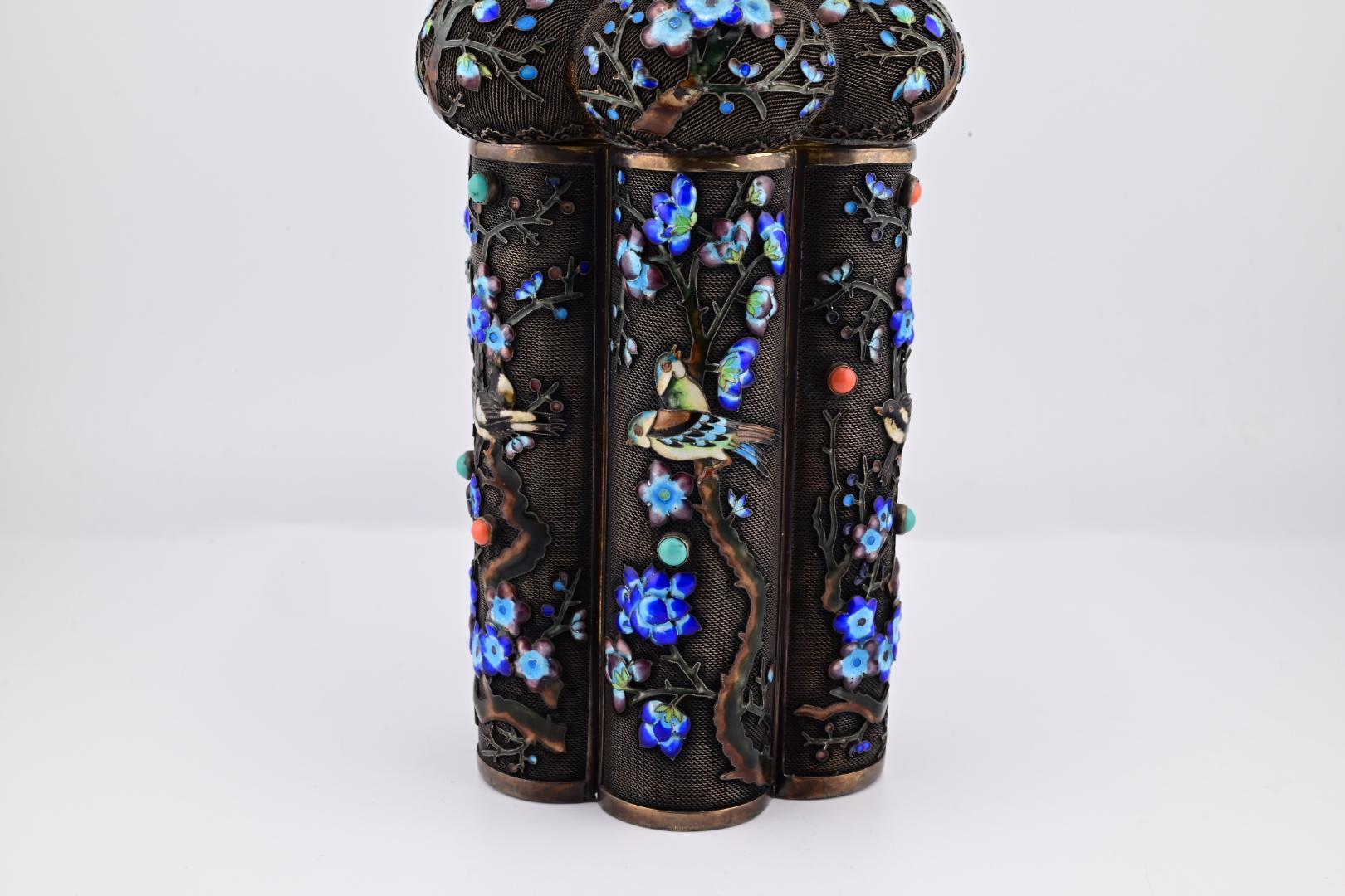 This is an intricately designed silver and enamel tea caddy. The piece features detailed floral and bird motifs in vibrant blue and multicolored enamel. The structure is cylindrical with a domed top, adorned with ornate patterns. The craftsmanship